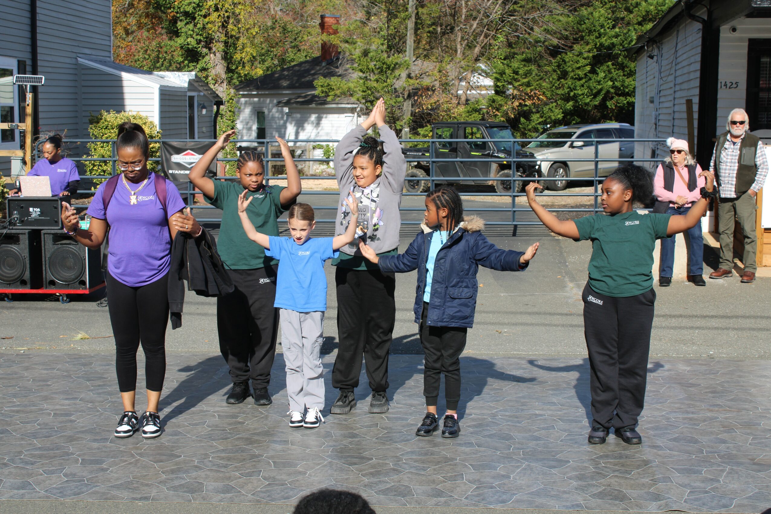 Group of children and adults holding hands and dancing outdoors on a paved area, with onlookers and trees in background.