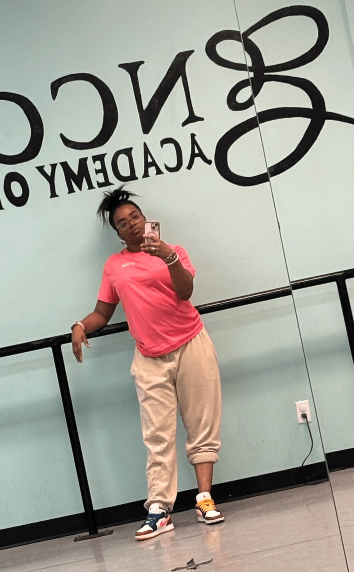 Person taking a mirror selfie in a dance studio, wearing a pink shirt and beige pants, with a large logo on the wall behind.