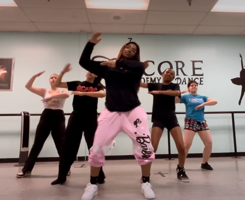 Five people dancing in a studio with a ballet barre, some with arms raised, one in pink pants and black top, others in casual clothing.