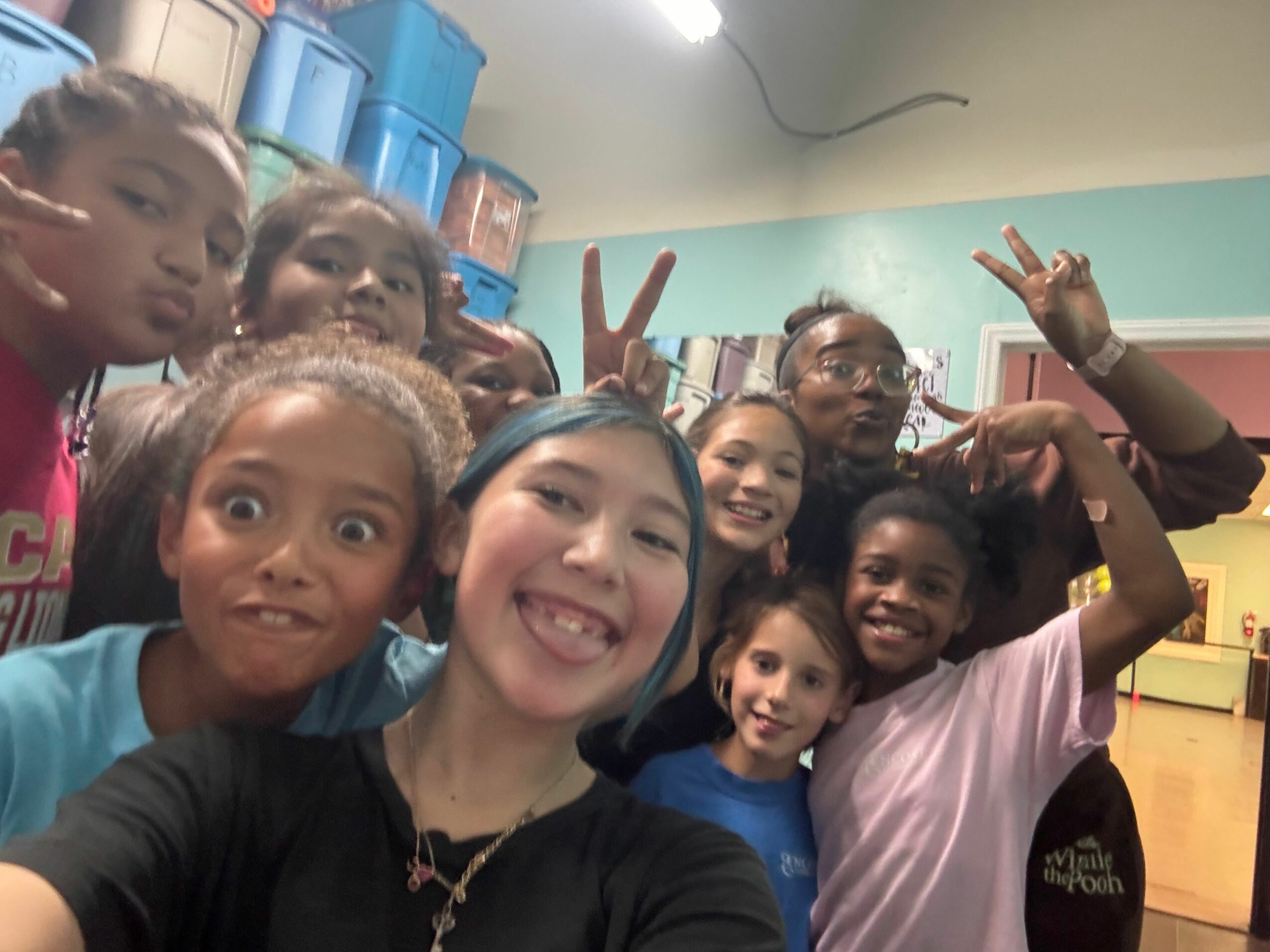Group of children and a woman smiling and making peace signs, indoors with colorful background.