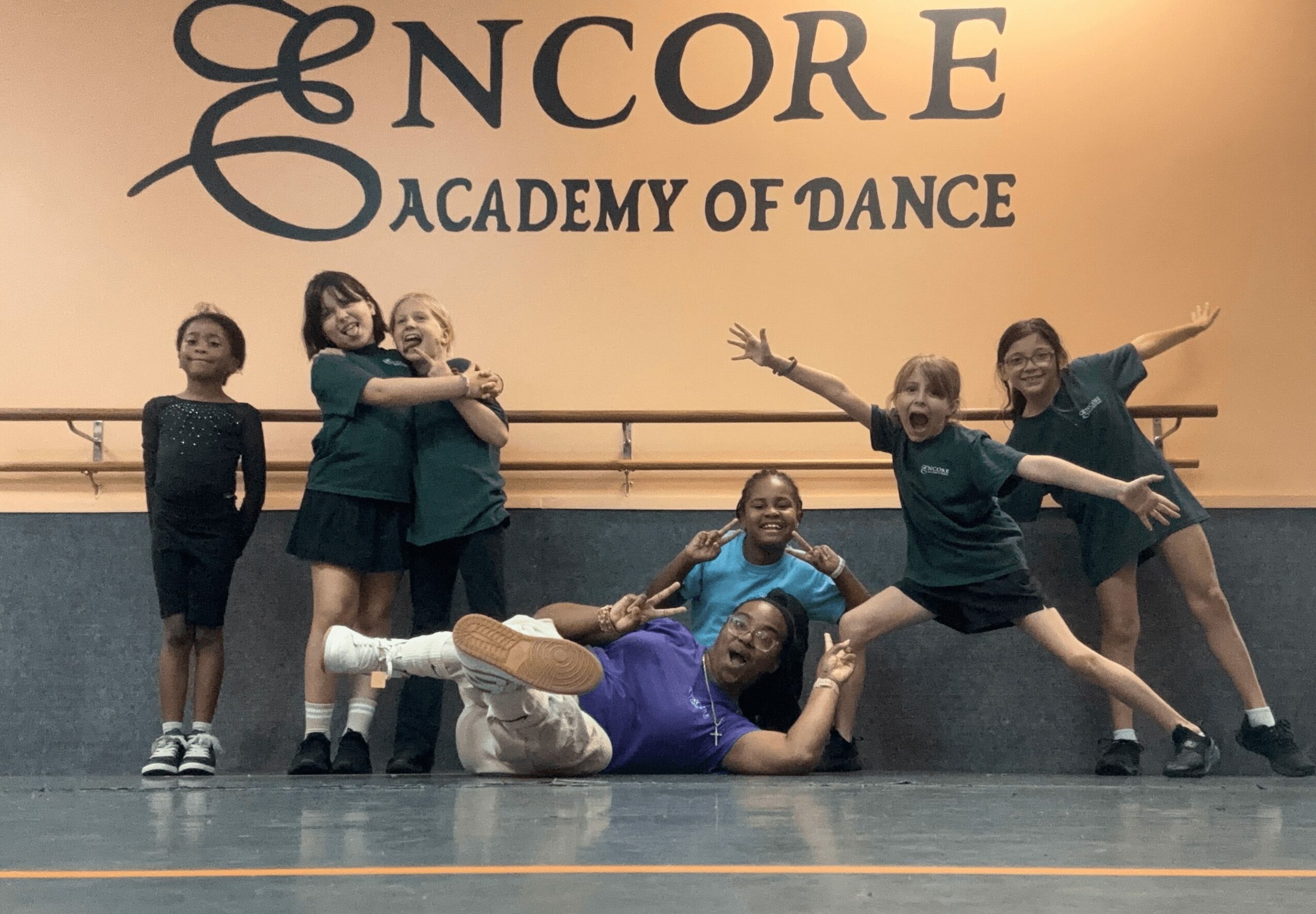 Group of children and an adult posing on dance studio floor with 'Encore Academy of Dance' sign behind them.