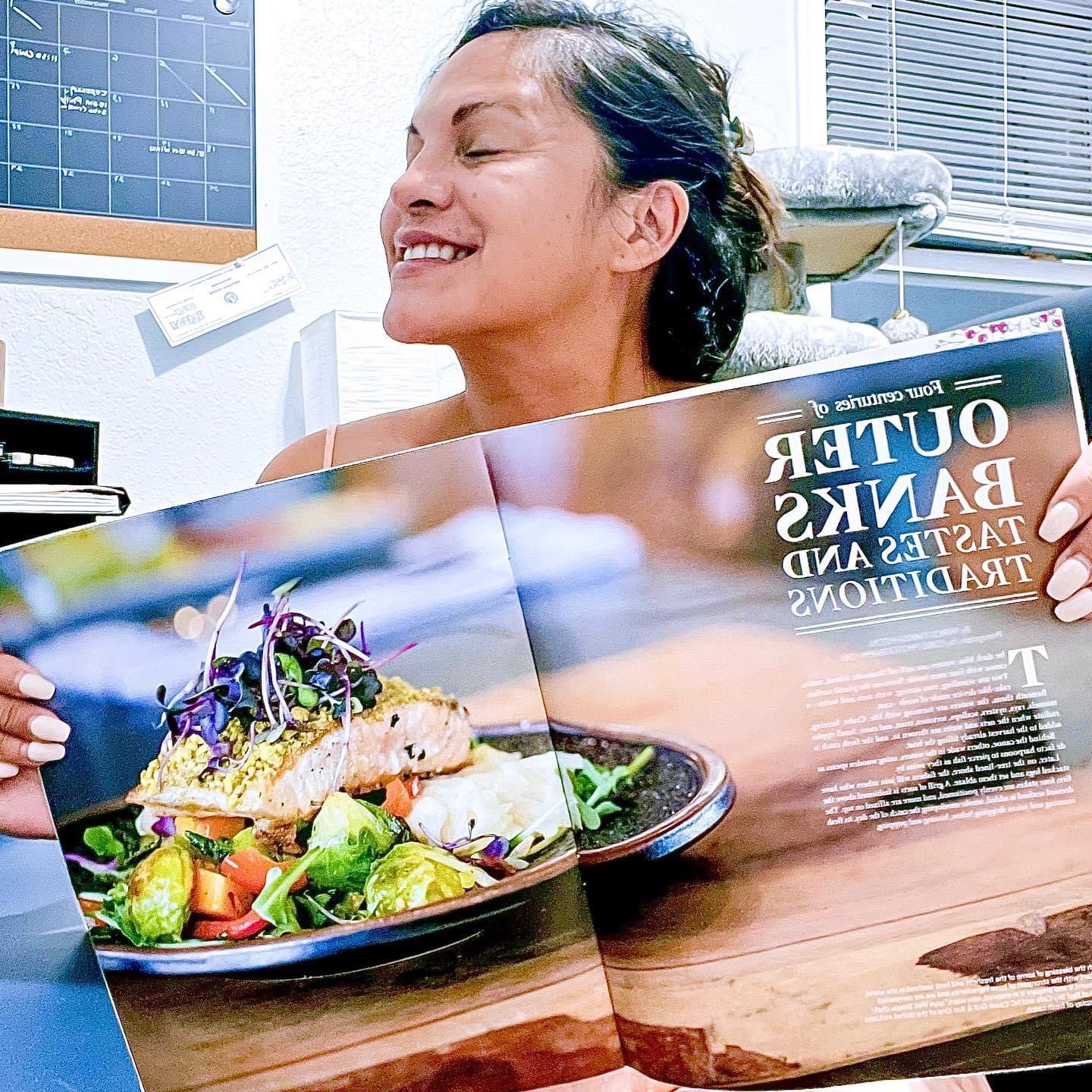 Smiling woman in a medical setting looking at a magazine with a plate of food on it.