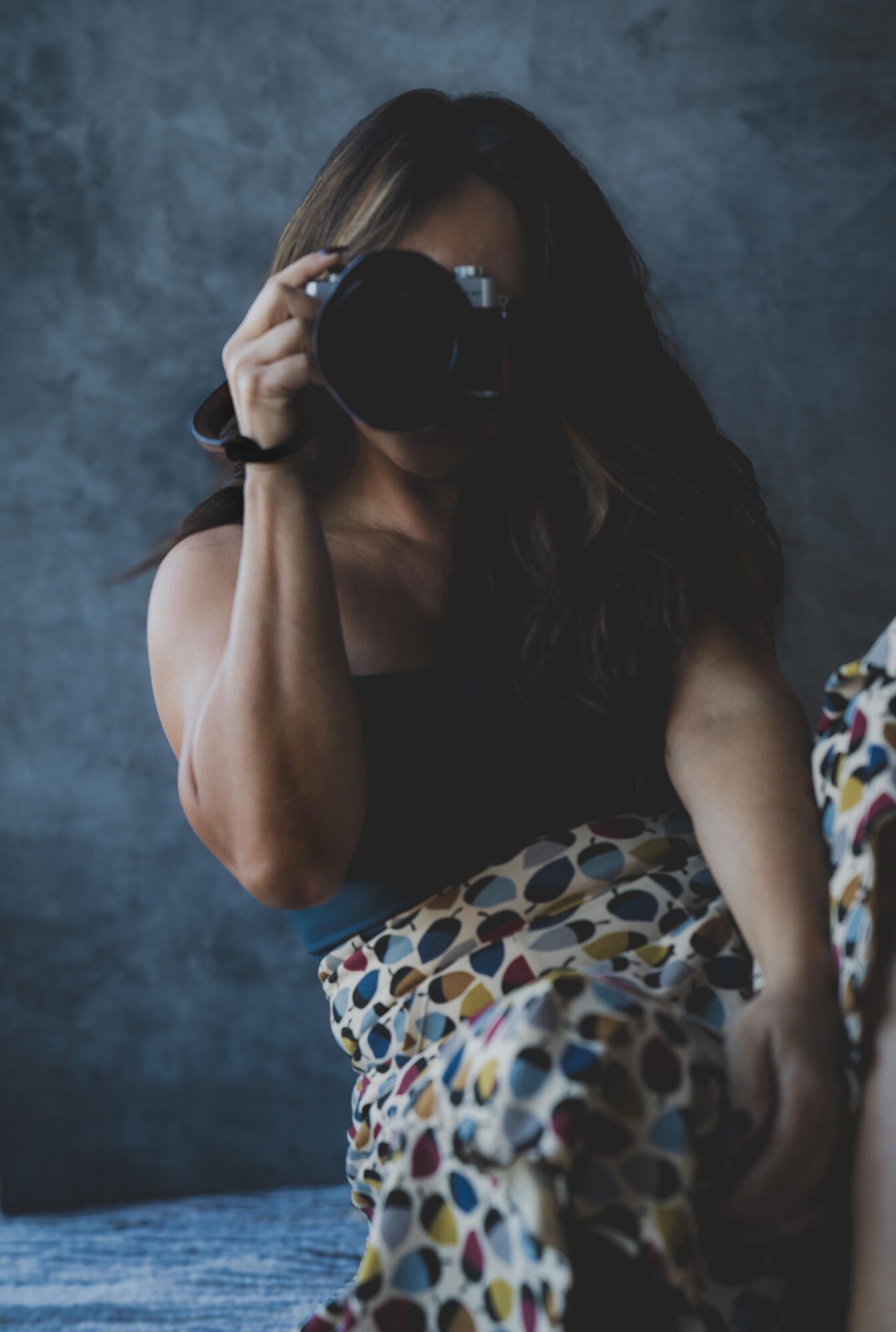 Person with long dark hair holding a camera, wearing a black top and patterned skirt, standing against a dark background.