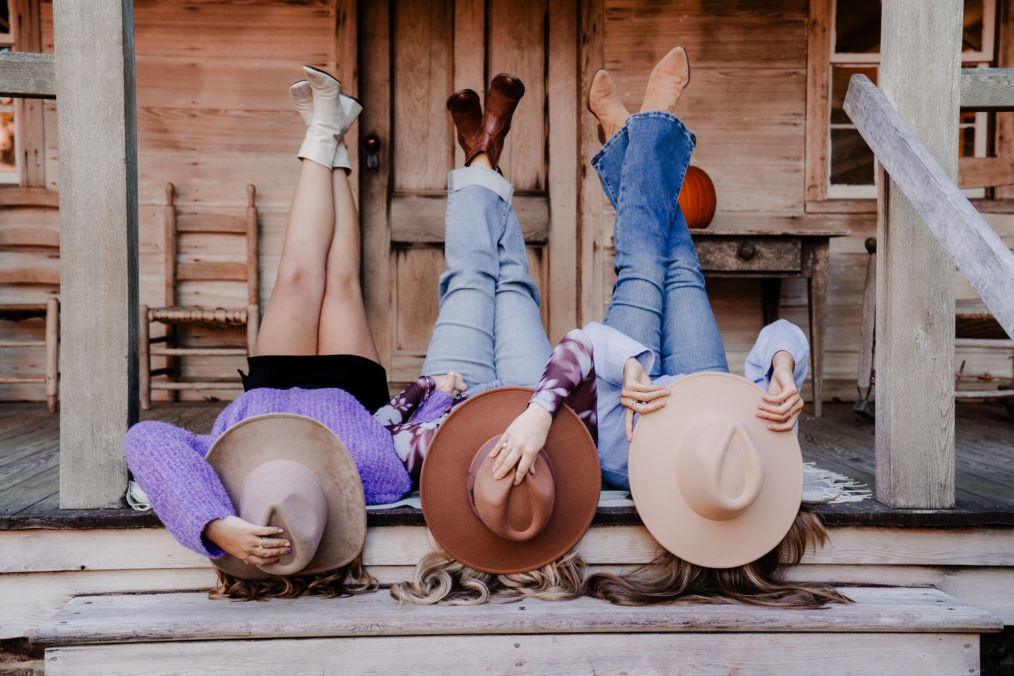 Three children lying on their backs on a wooden porch, holding hats over their faces, with a wooden wall and chairs behind them.