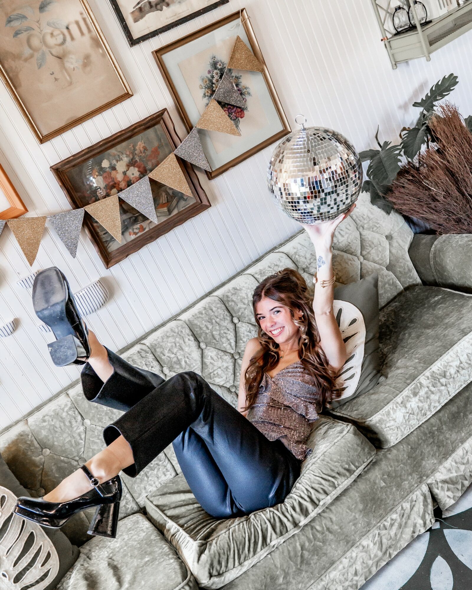 Young woman lying on a sofa, holding a disco ball, smiling, with decorative wall art and plants in the background.