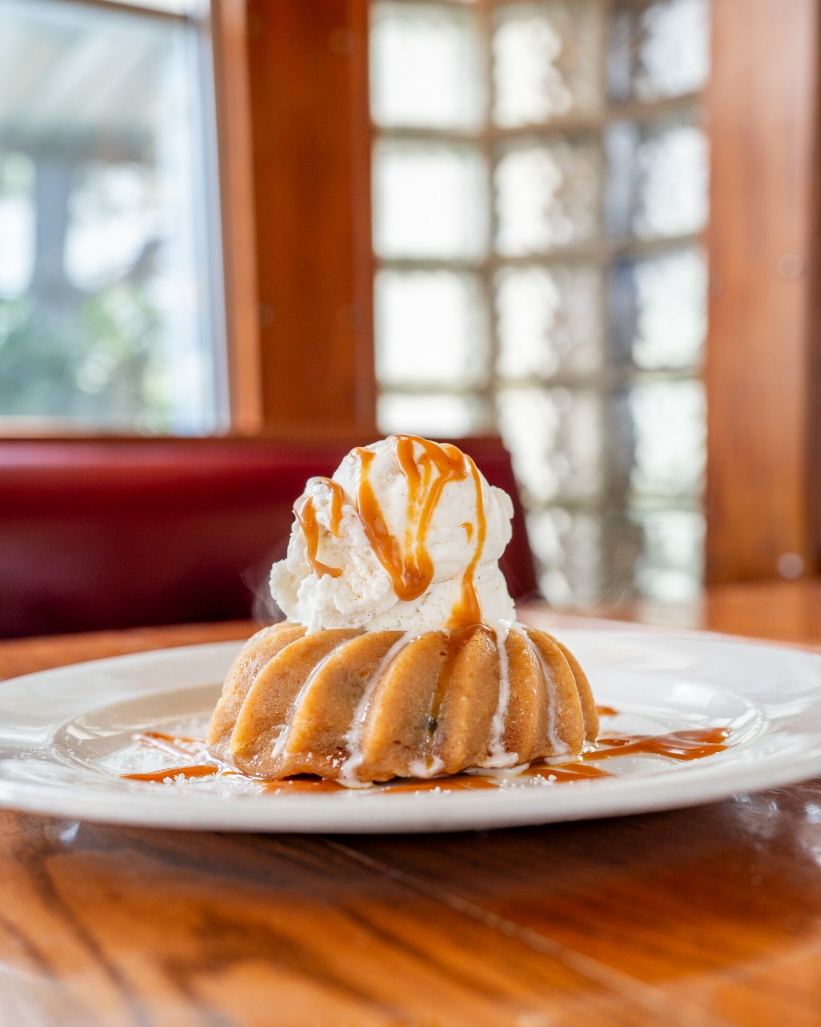 A dessert with a scoop of ice cream topped with caramel sauce on a plate with a bundt cake, set on a wooden table.