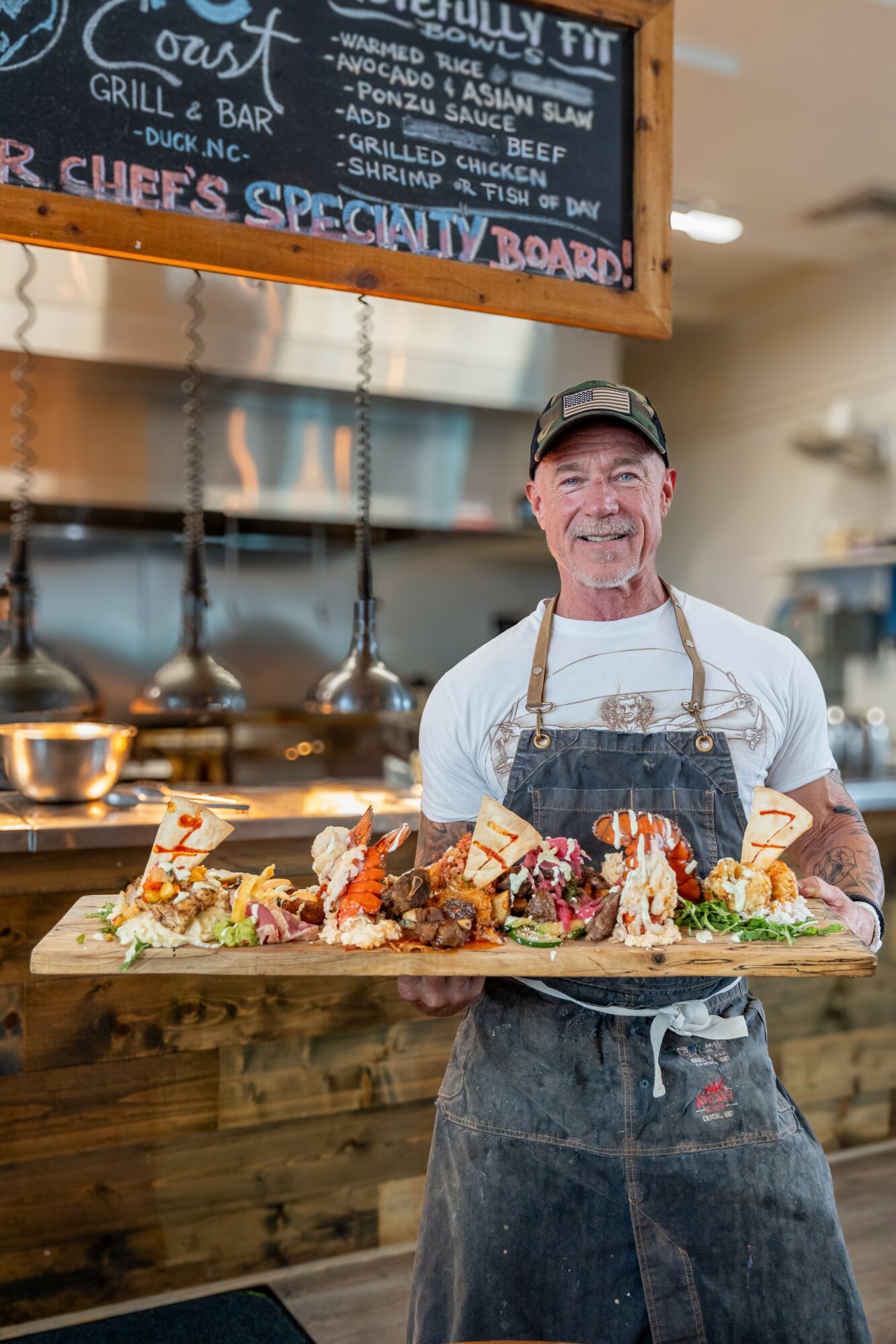Smiling man wearing apron and cap stands behind a wooden counter with a large tray of food, in a restaurant kitchen.