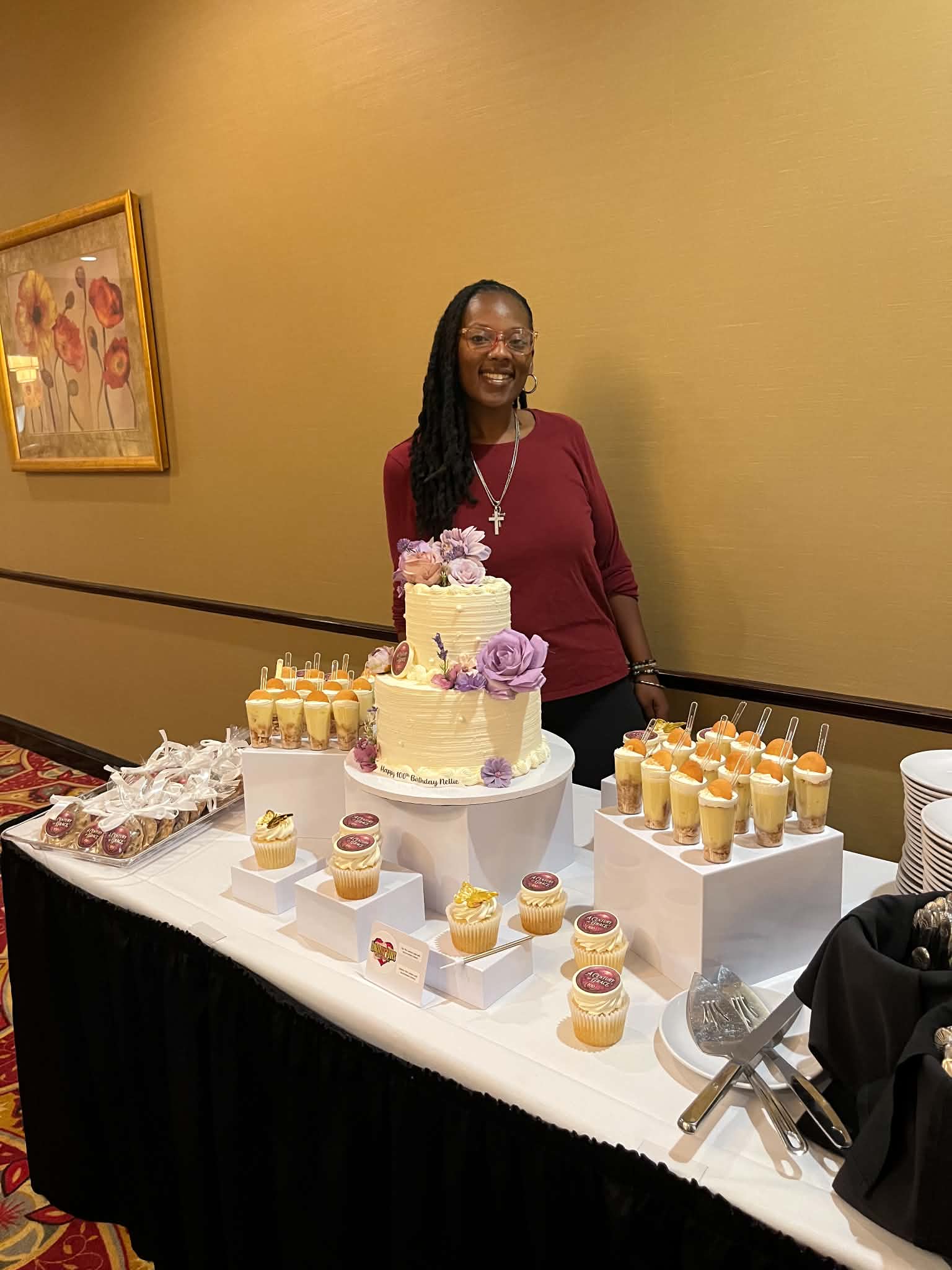 Woman with long braids and glasses smiling behind a decorated cake table, yellow wall, framed artwork, birthday celebration.