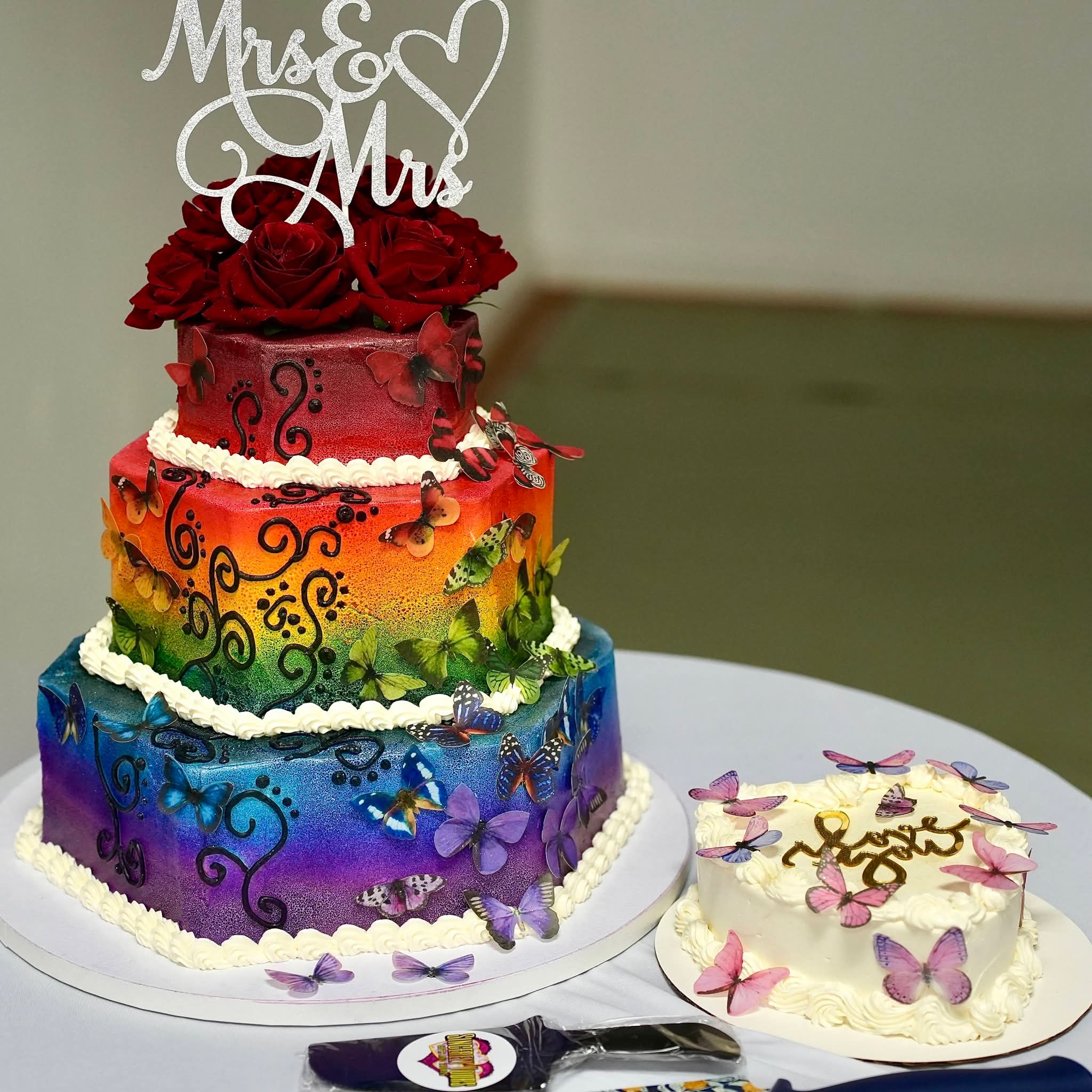 Colorful tiered wedding cake with red roses and a topper reading 'Mrs & Mrs', next to a white cake with purple butterflies.