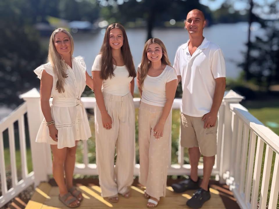 Four people standing on a porch railing outdoors, smiling, with trees and water in the background.