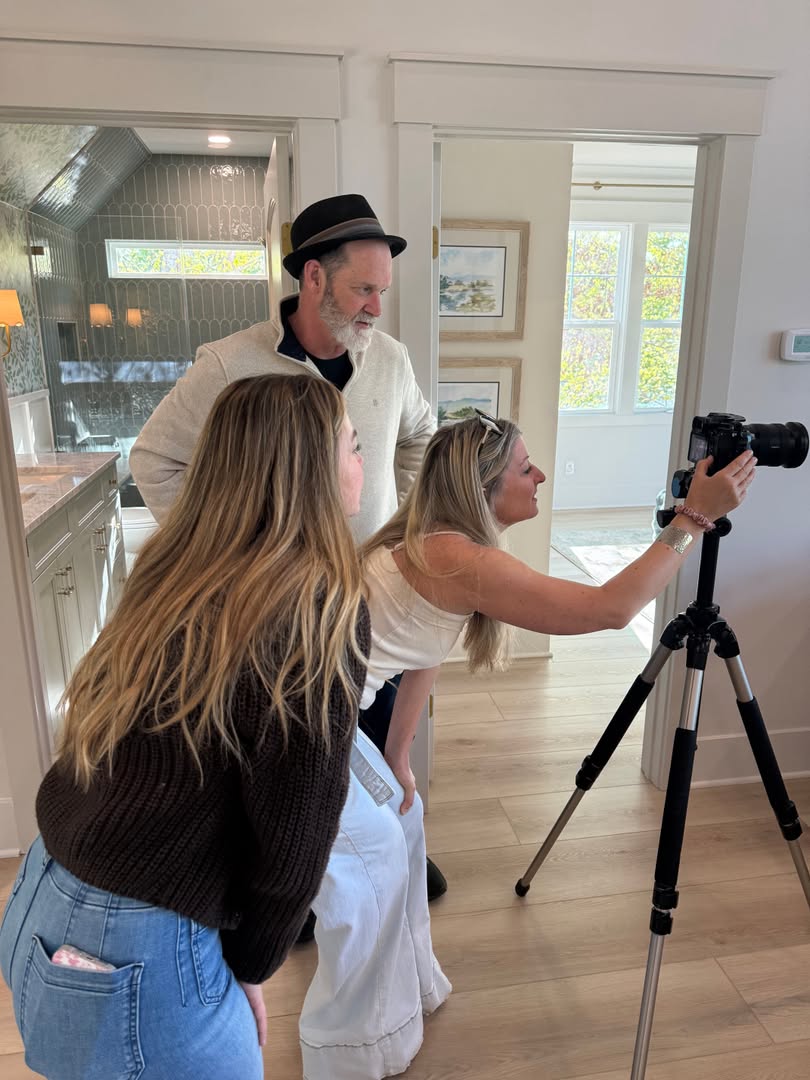 Three people in a bright room, one woman taking a photo with a camera on a tripod, two others watching, natural light from windows.