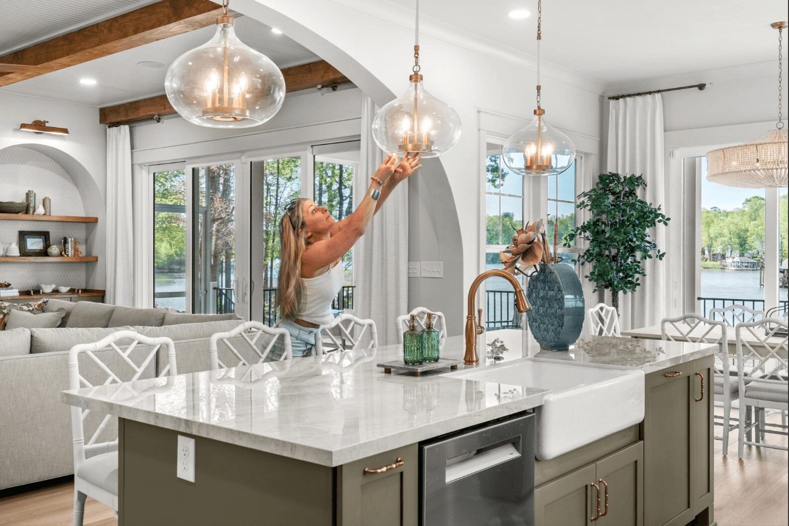 Woman adjusting pendant lights in a bright, modern kitchen with a large island, white cabinets, and large windows.