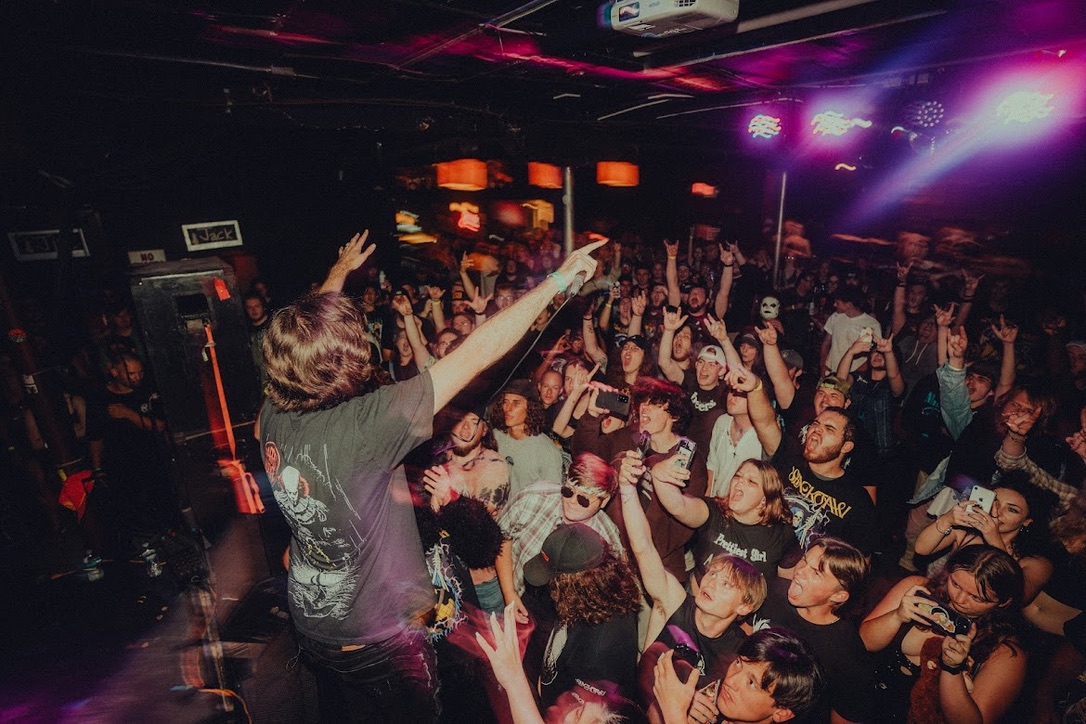 Person on stage with arm raised performing in front of a crowd at a nightclub, colorful lights overhead, lively atmosphere.