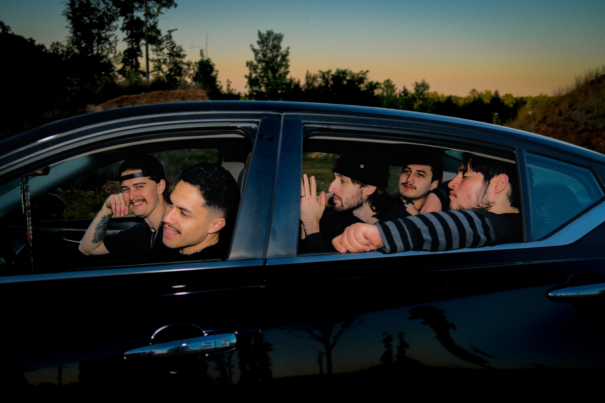 Five people sitting in a car during sunset, smiling and talking, with trees and sky in the background.