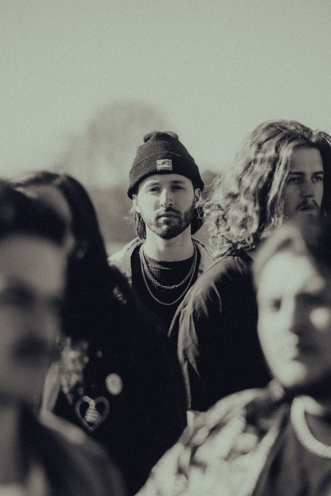 A young man with a beard, wearing a beanie and layered necklaces, stands among a group of people outdoors.