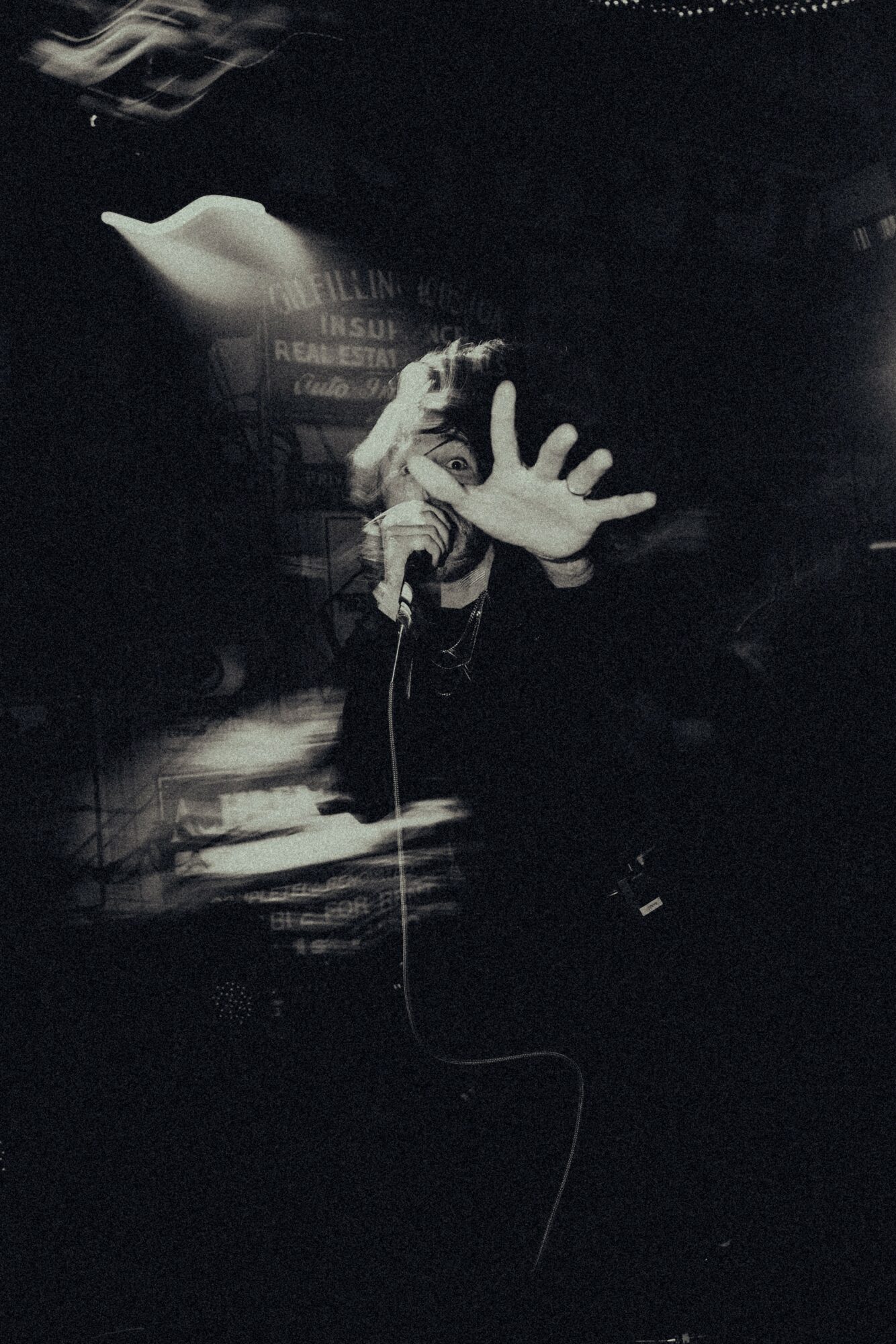 Person on stage reaching out with hand, holding a microphone, in a dark setting with stage lights.