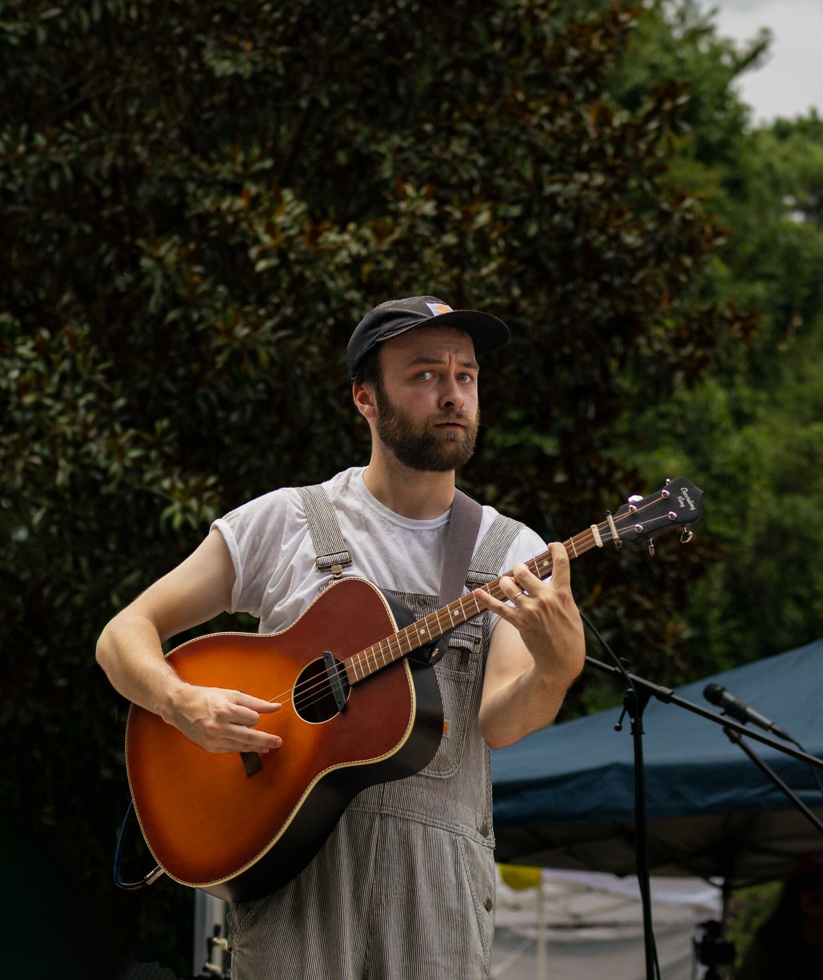 Man playing guitar outdoors with trees and a tent in the background.