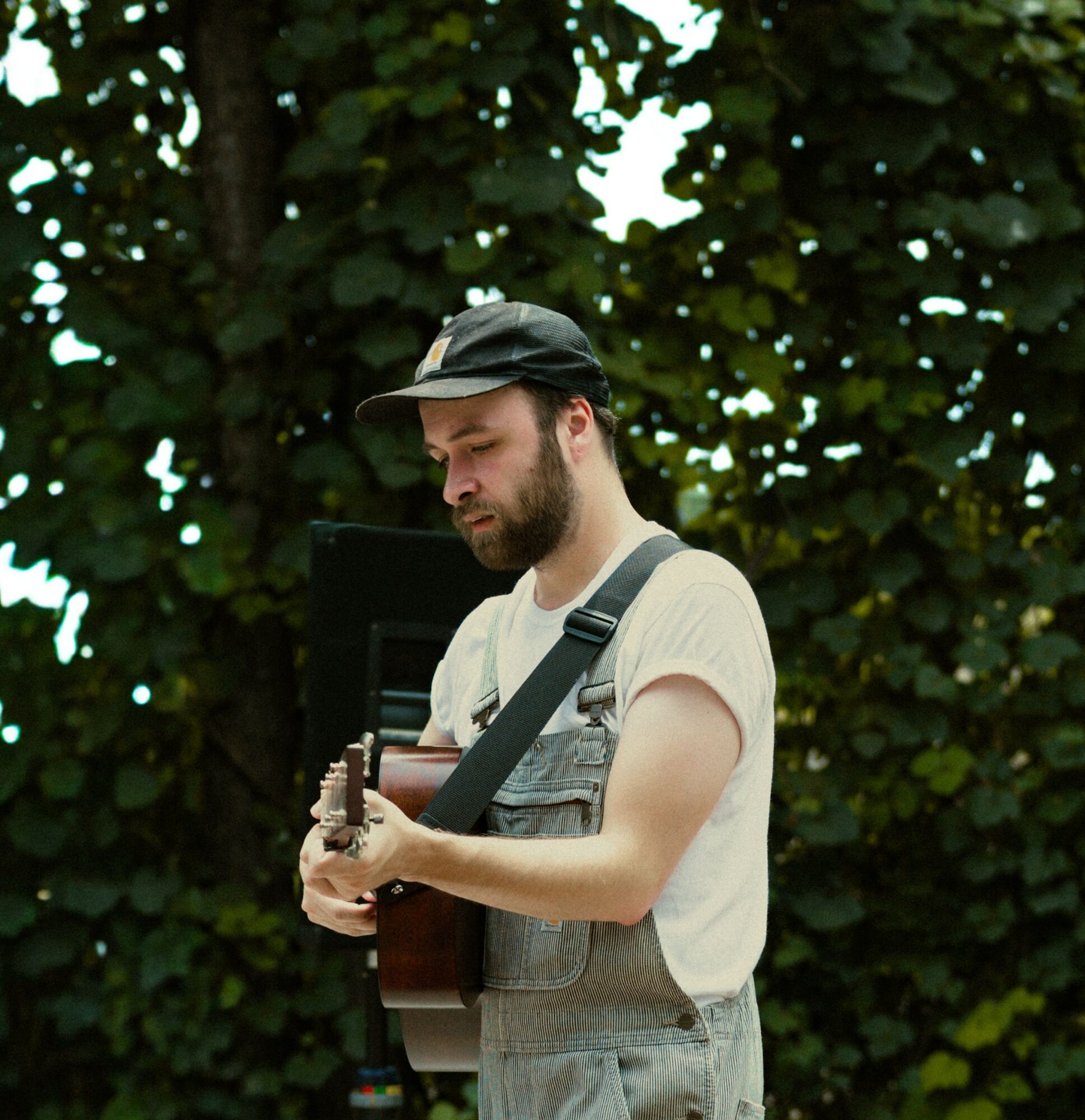 Young man with a beard playing an acoustic guitar outdoors, wearing a cap, white t-shirt, and overalls, standing in front of leafy background.