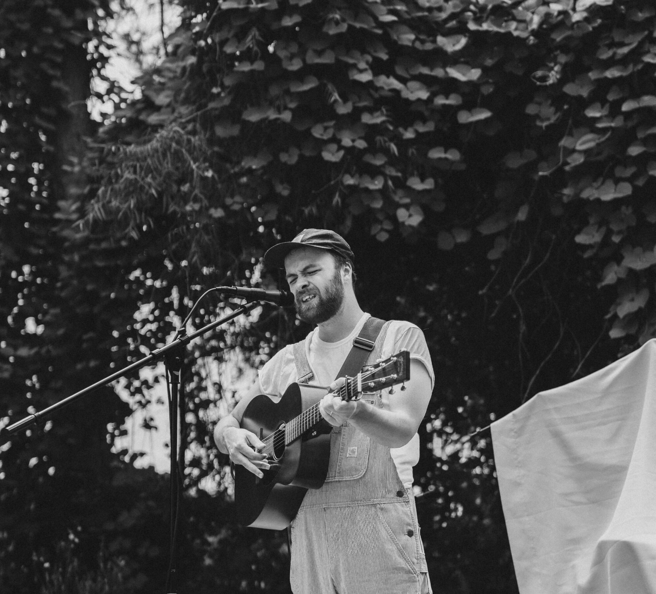 Person playing guitar and singing outdoors with trees in background, smiling, wearing a hat and light-colored clothing.