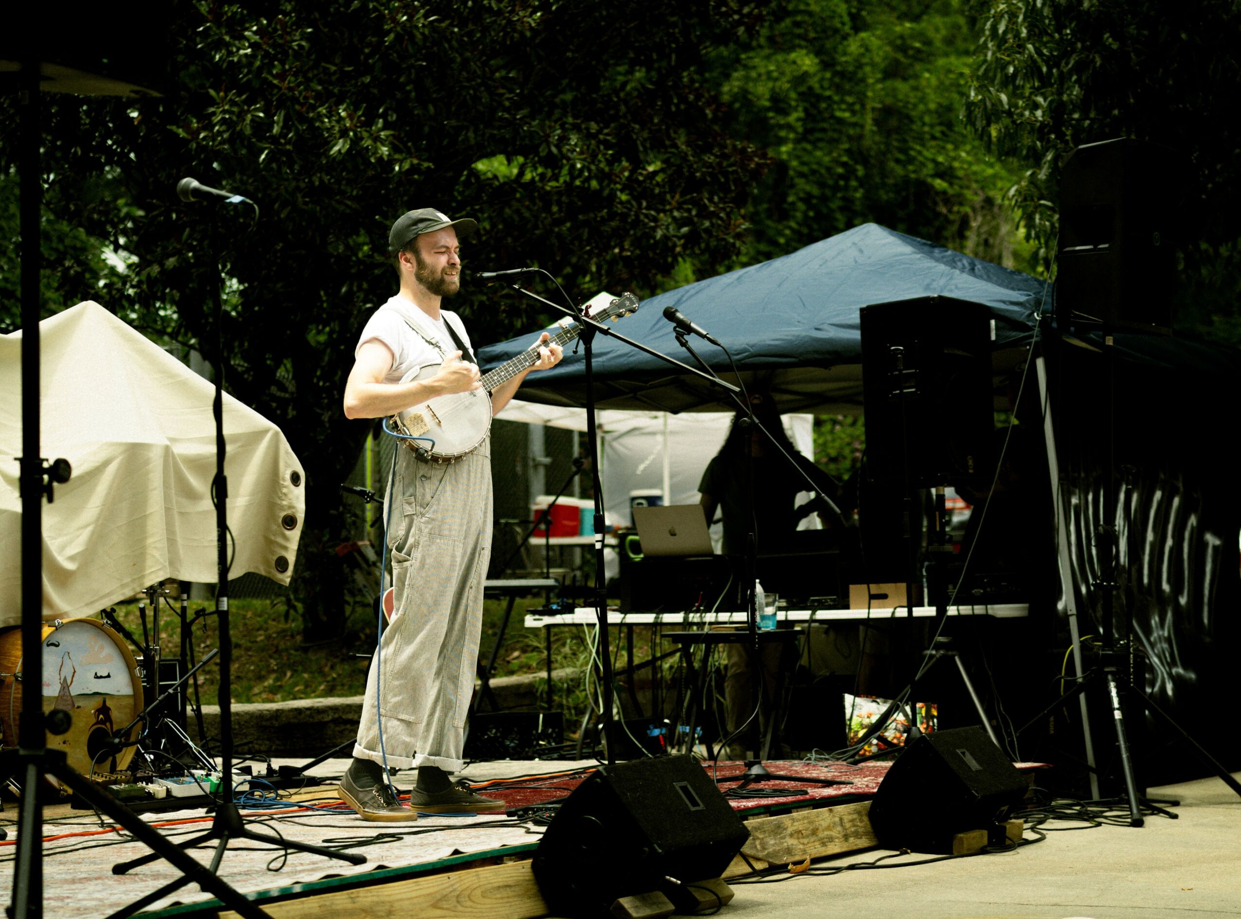 Man playing banjo on outdoor stage with microphones, speakers, and tents in background.