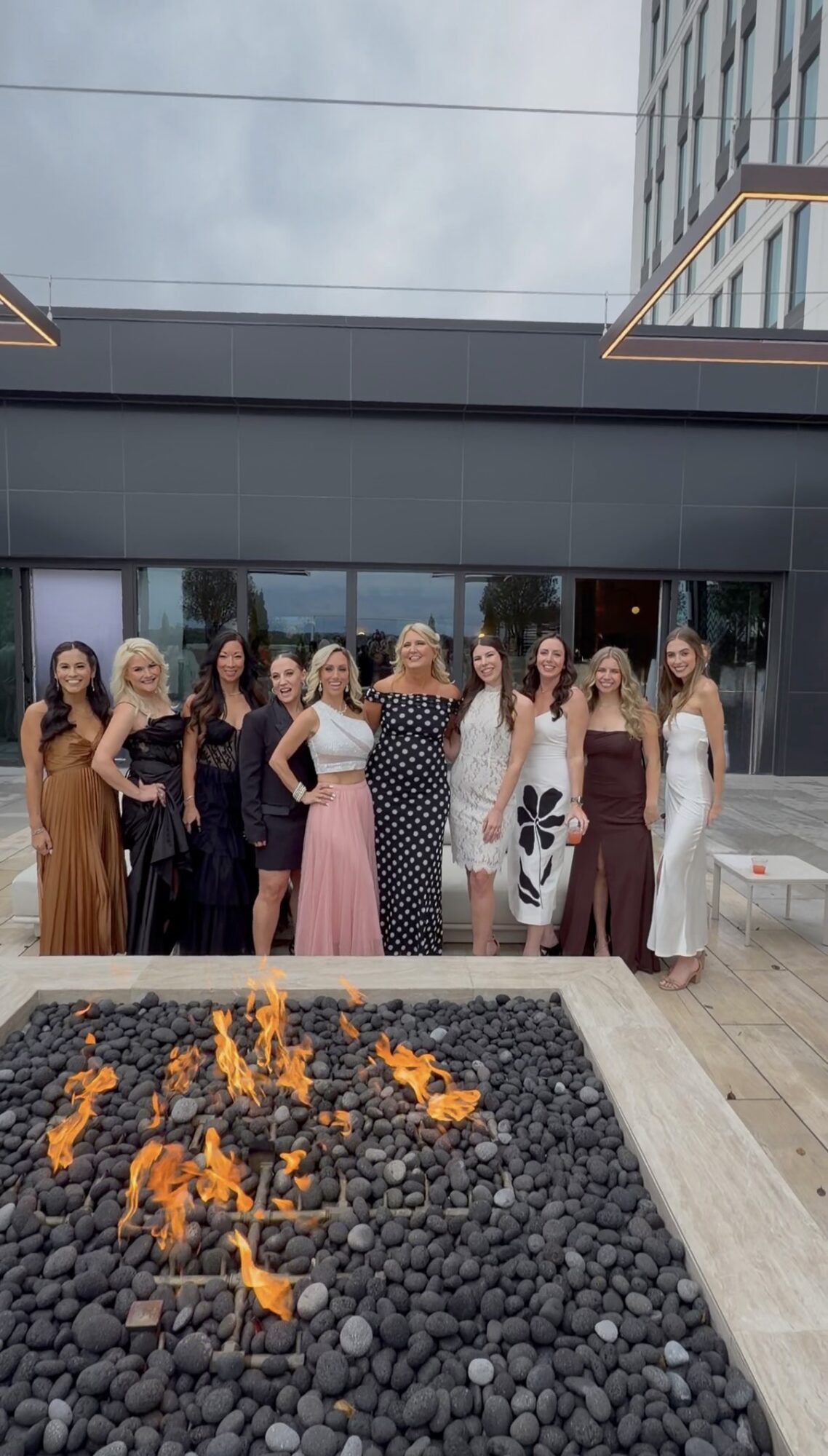 Group of ten women standing outdoors in front of a modern building, with a fire pit in the foreground.