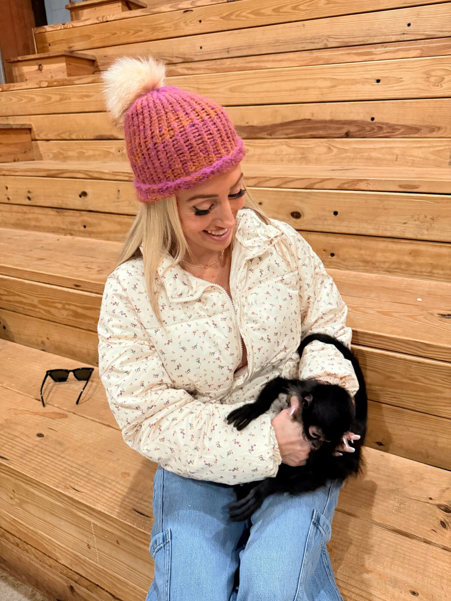 Person lying on wooden floor holding a small black and brown dog, wearing a pink knit hat with a pom-pom, white sweater, and blue jeans.