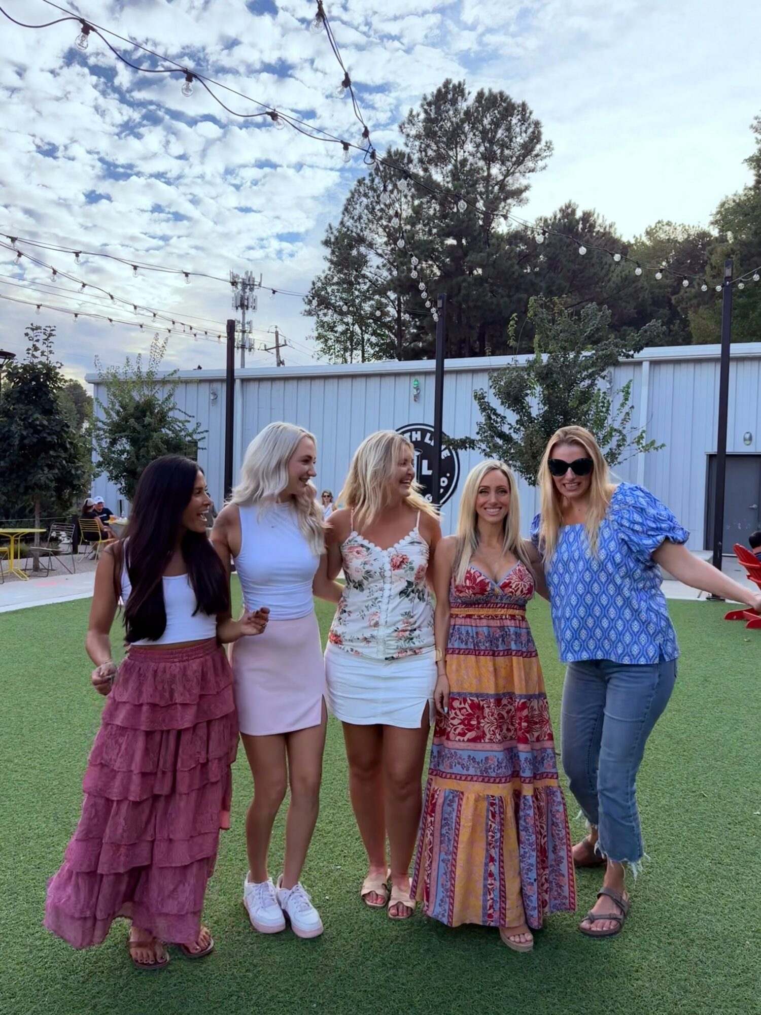 Five women standing outdoors on grass, smiling, with trees and a white fence in the background.