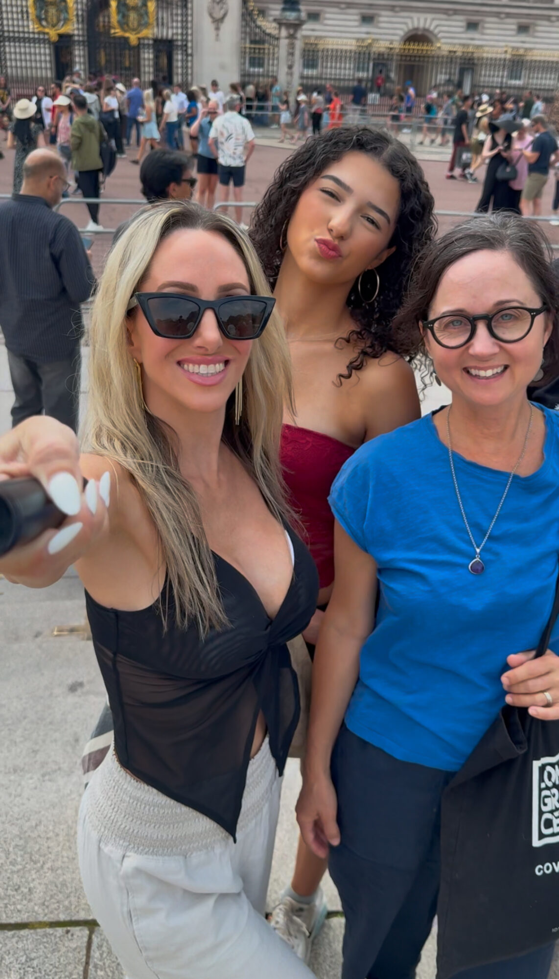 Three women smiling and posing for a selfie in a crowded outdoor area with historic buildings in the background.