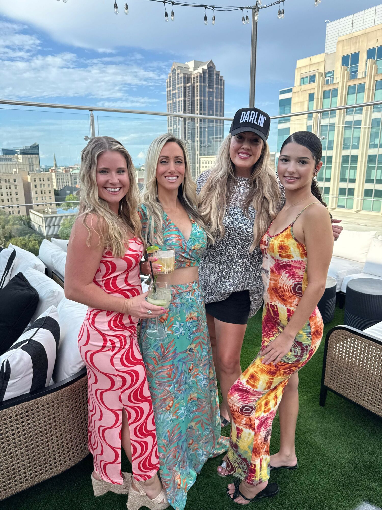 Four women standing outdoors on a rooftop with city buildings in the background, dressed in colorful summer outfits, smiling.