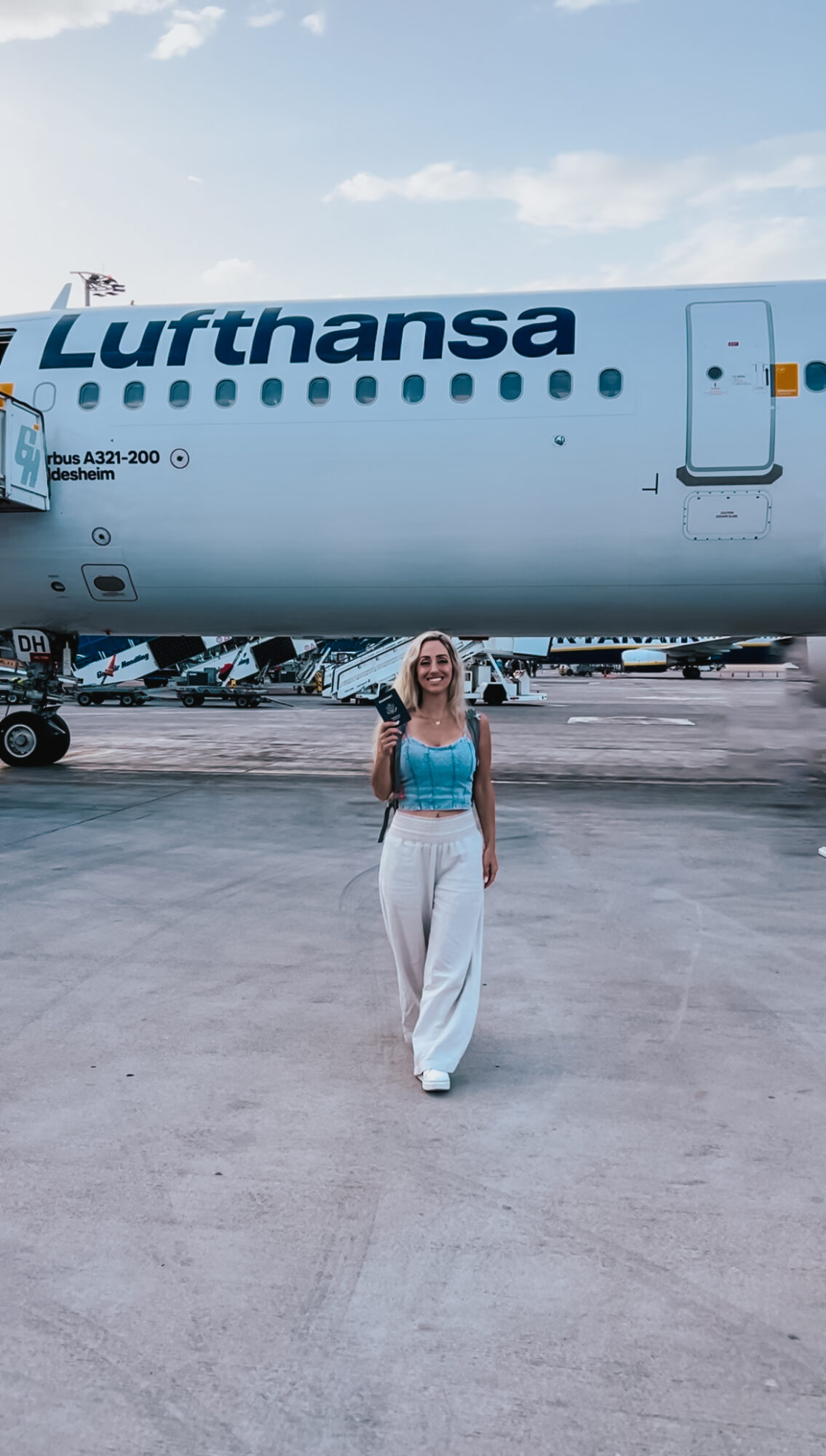 Woman standing on tarmac in front of Lufthansa airplane, smiling, wearing a blue top and white pants.