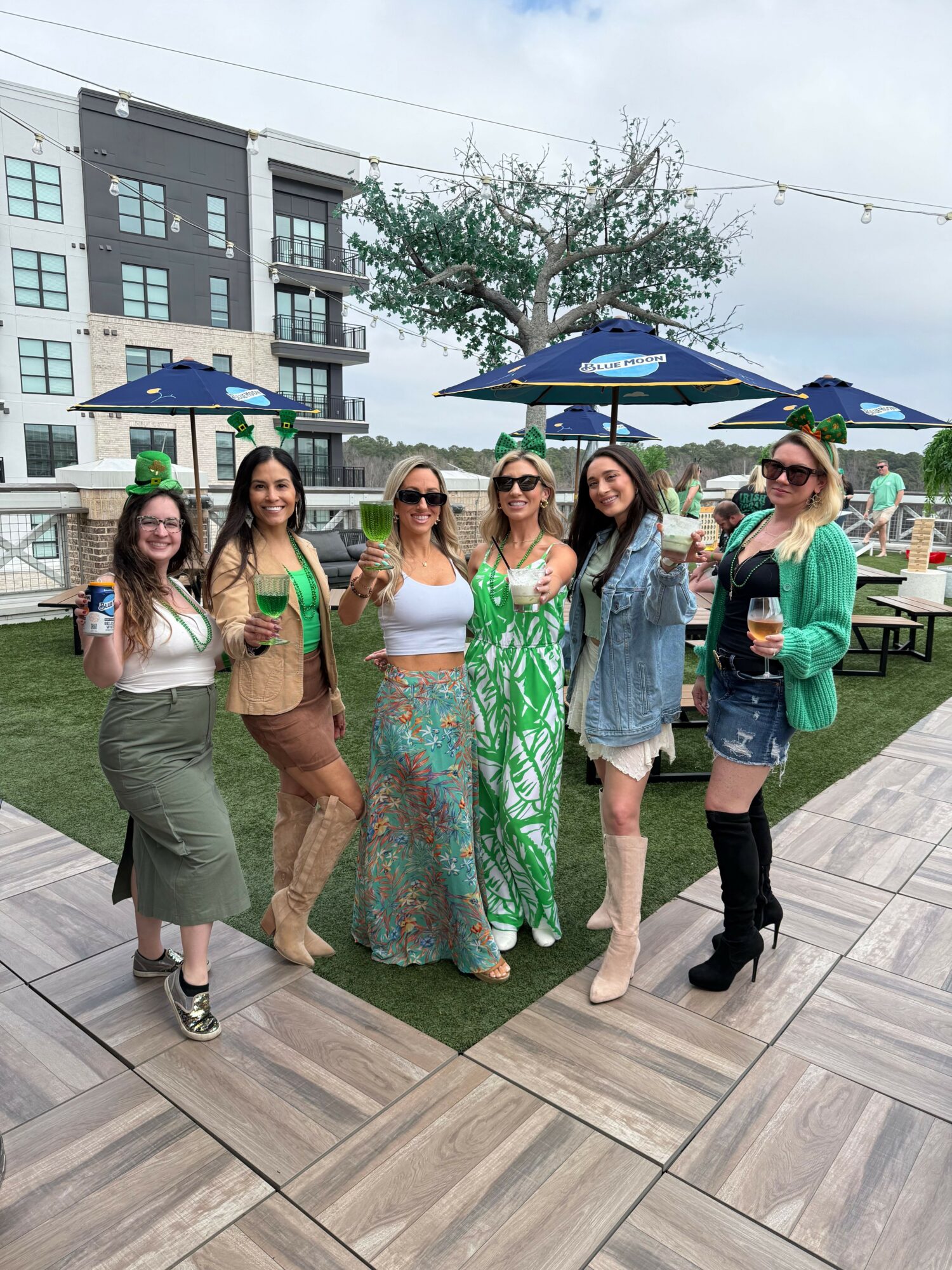 Group of six women standing outdoors on a wooden deck, with buildings and trees in the background, smiling and posing for a photo.