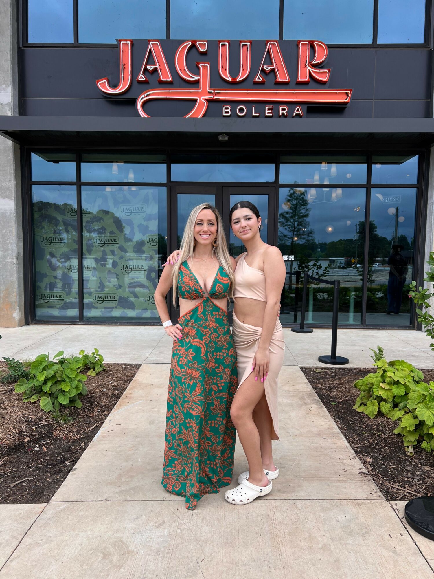 Two women standing outside a building with glass doors, one in a beige dress and the other in a green patterned dress, holding drinks.