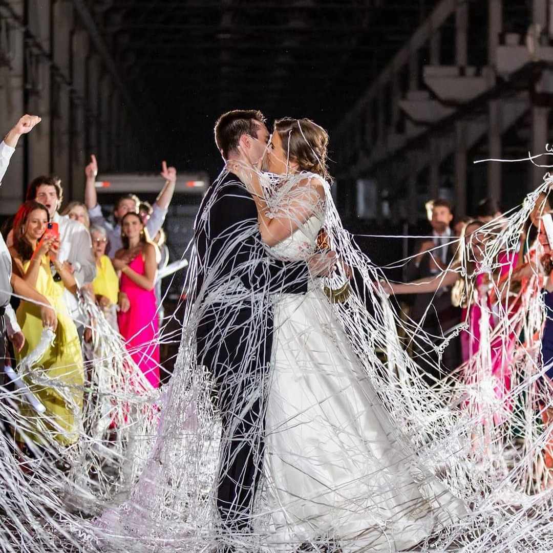 Couple in wedding attire kissing surrounded by guests throwing streamers, celebration in a large indoor venue.