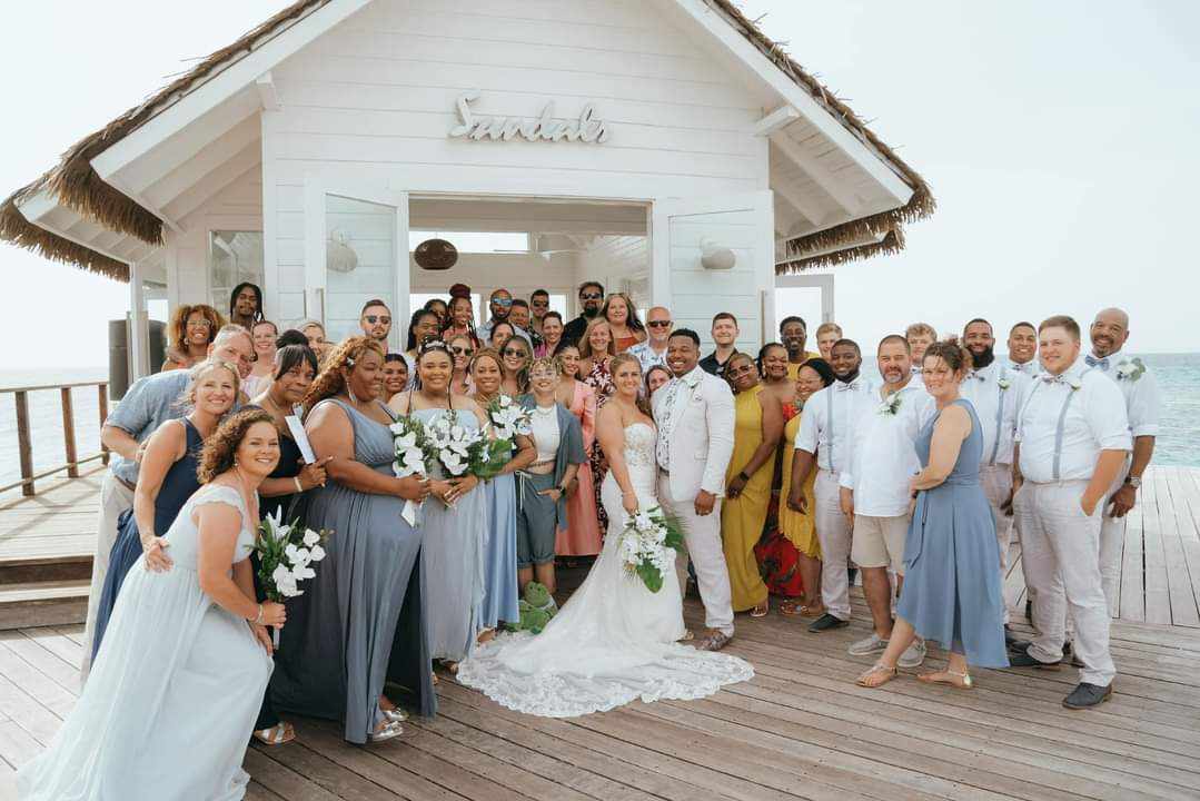 Group of people, including a bride and groom, gathered outside a white building with a thatched roof, celebrating a wedding.