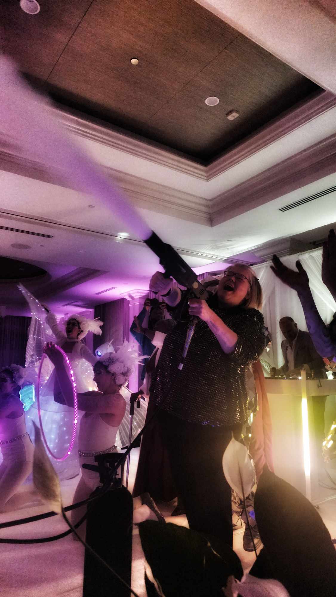 Person holding a fog machine emitting purple fog at an indoor event with colorful lighting and people in the background.