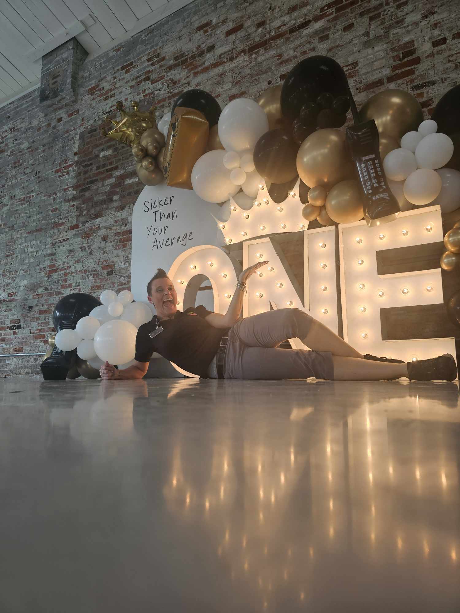 Person lying on the floor near large illuminated 'LOVE' sign with black, white, and gold balloons, brick wall background.