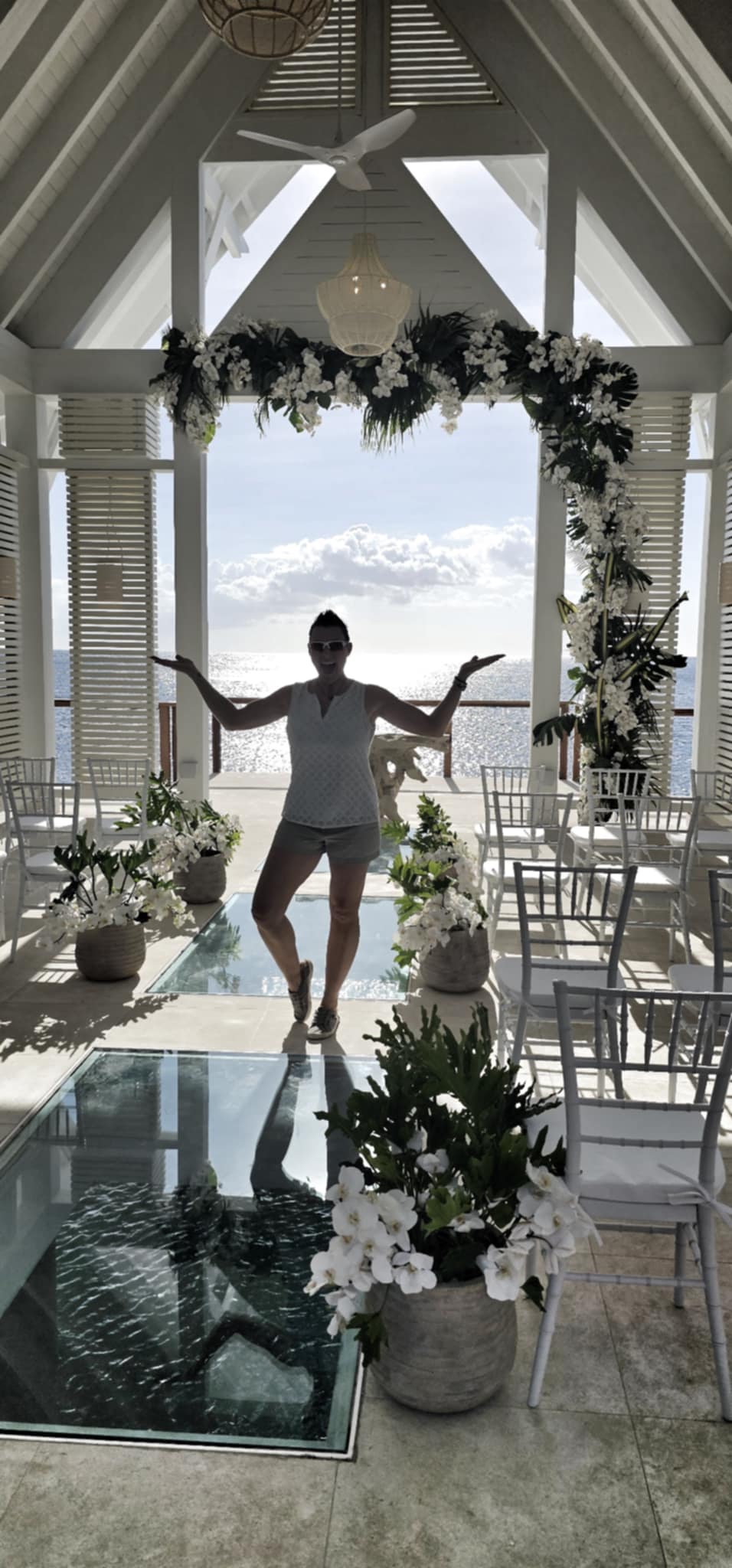 Person standing with arms outstretched in a decorated indoor space with large windows and a view of the sky.