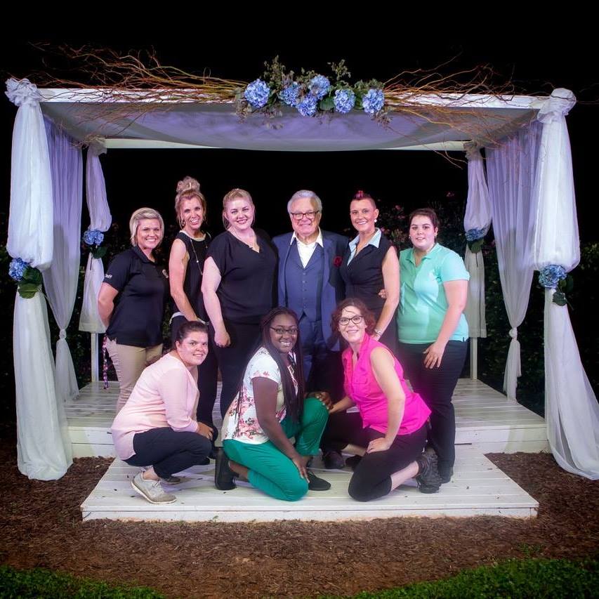 Group of nine people posing under a decorated outdoor canopy with white drapes and flowers, on a wooden platform.
