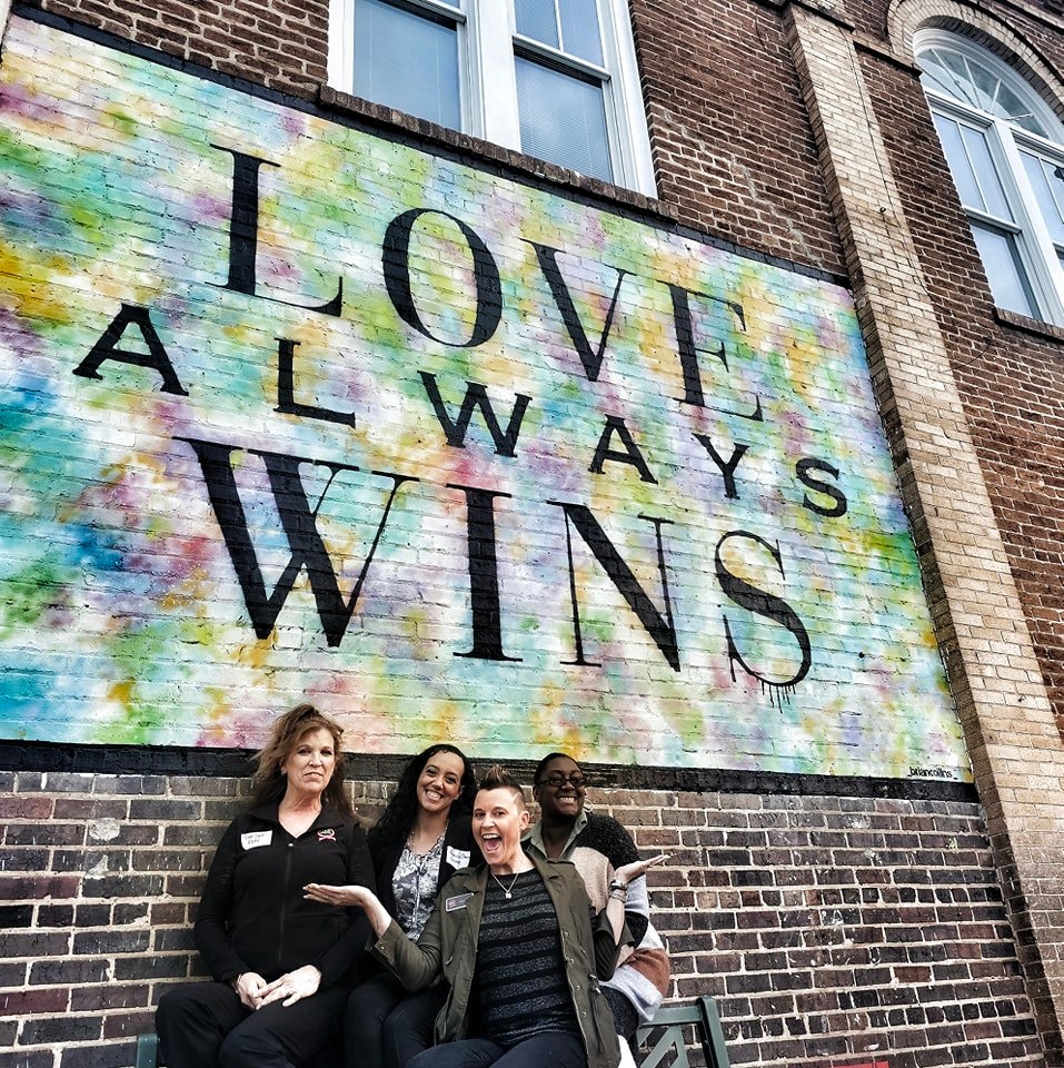 Three women standing in front of a colorful mural with the words 'LOVE ALWAYS WINS' on a brick wall.