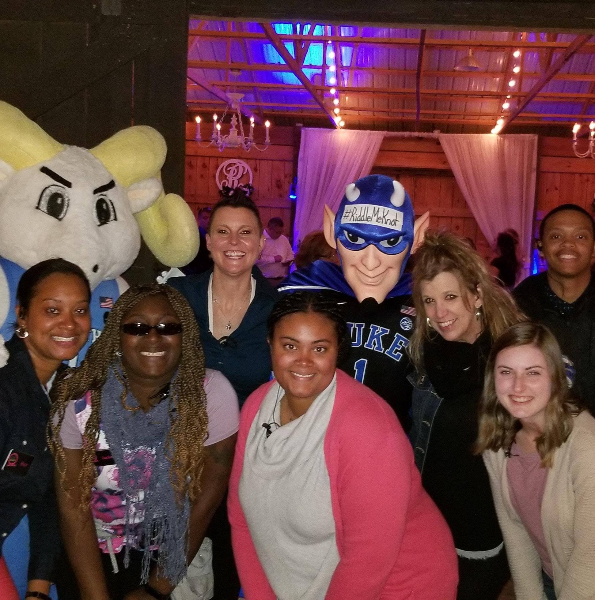 Group of smiling people with a mascot in a decorated indoor setting, some wearing sunglasses and casual clothing.