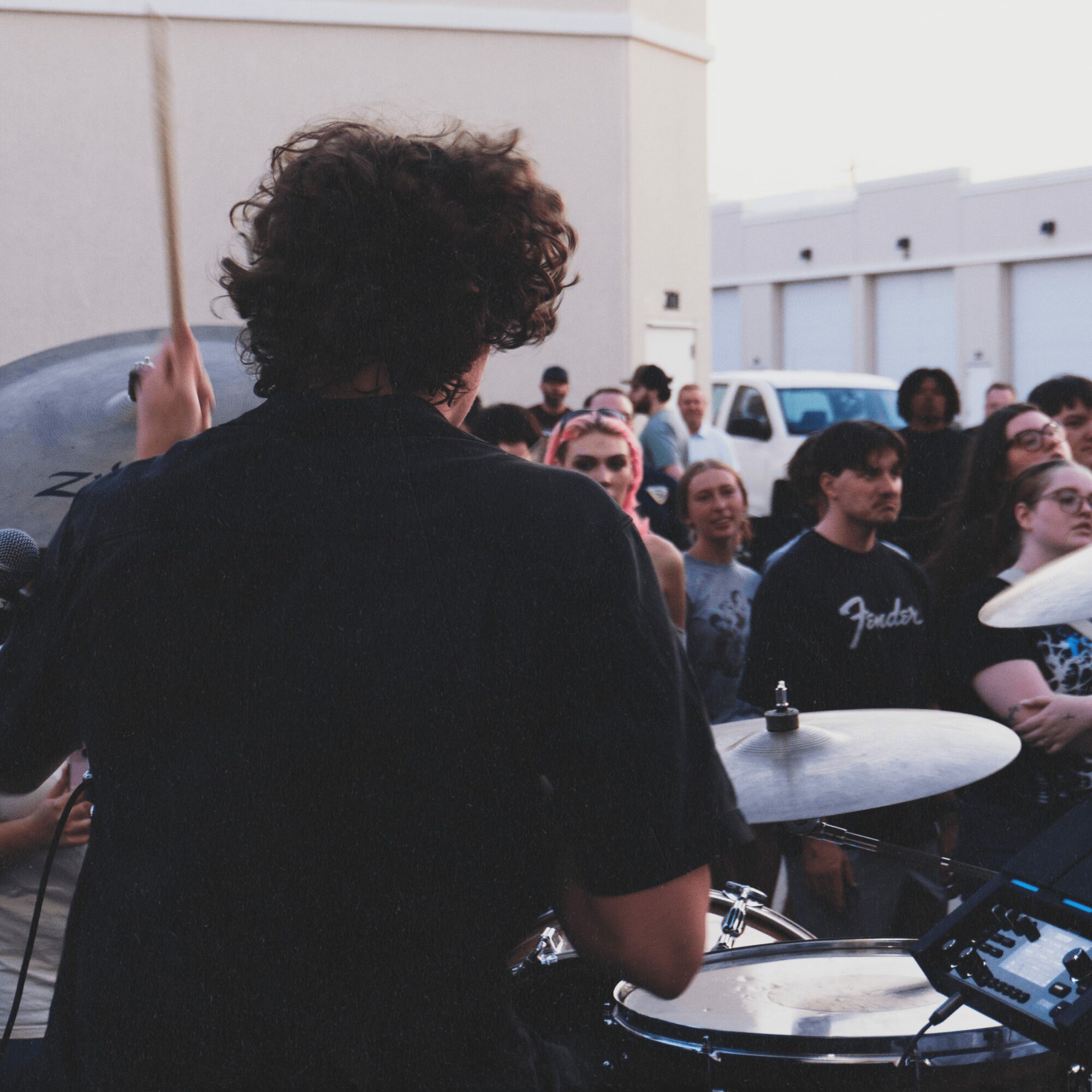 Person with curly hair playing drums at an outdoor event, with a crowd of people in the background.