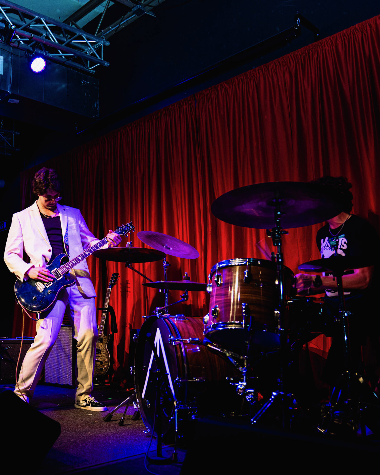 Two musicians perform on stage with red curtain backdrop, one playing guitar and the other on drums, stage lighting visible.