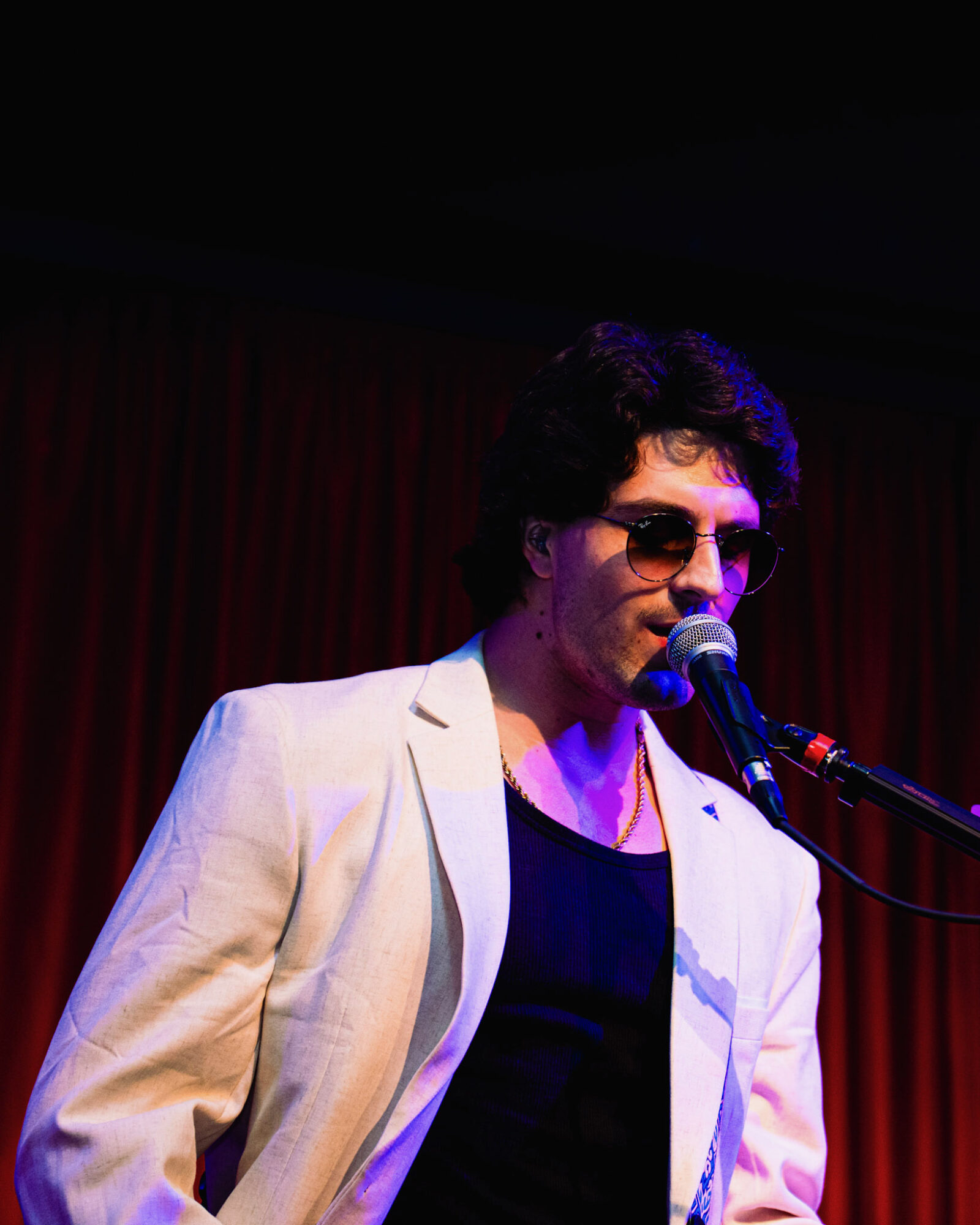 Person with dark curly hair wearing sunglasses and a light-colored blazer, singing into a microphone against a red curtain background.