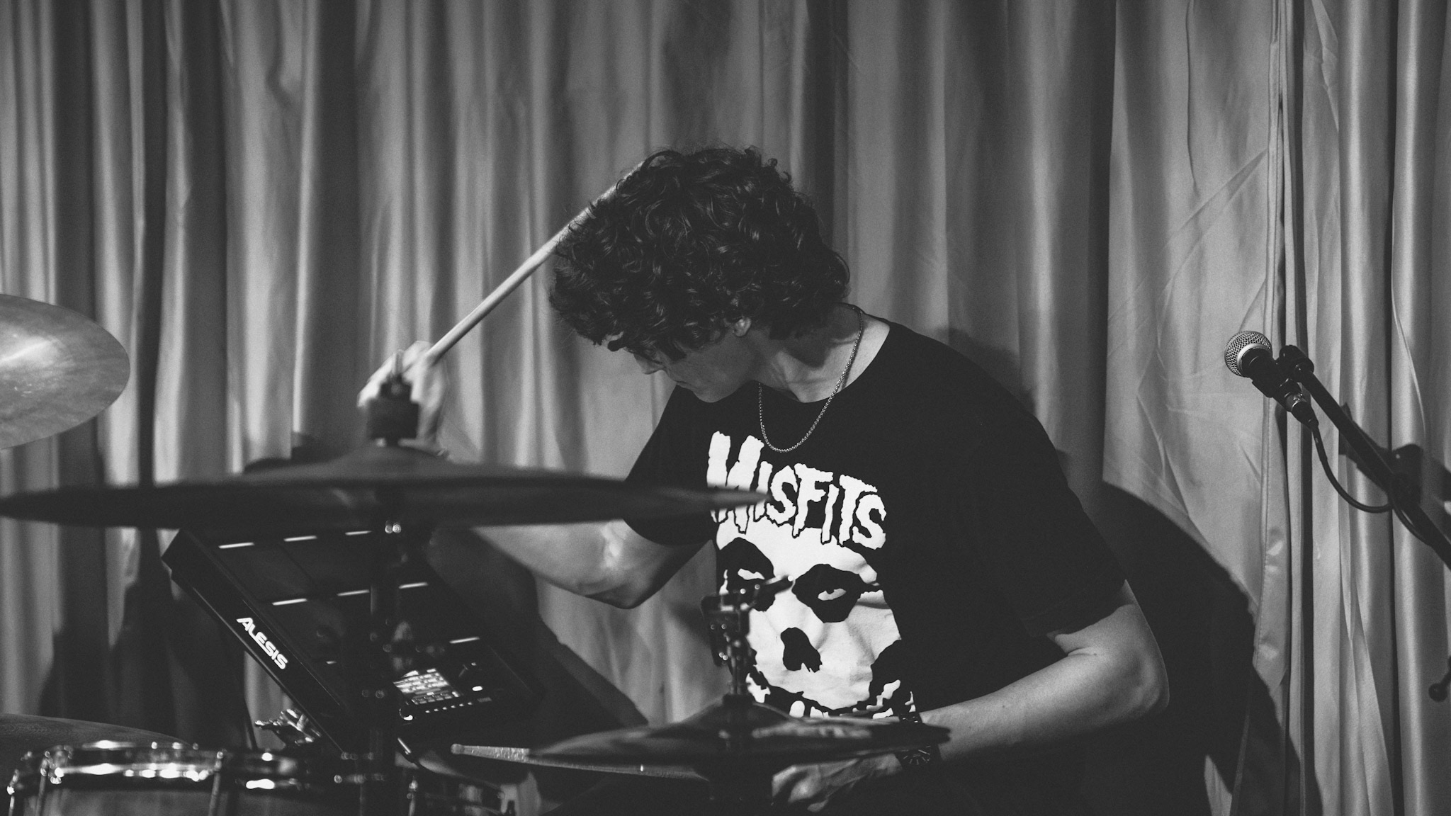 Person with curly hair playing electronic drum set in front of curtain, wearing a graphic T-shirt, with microphone nearby.