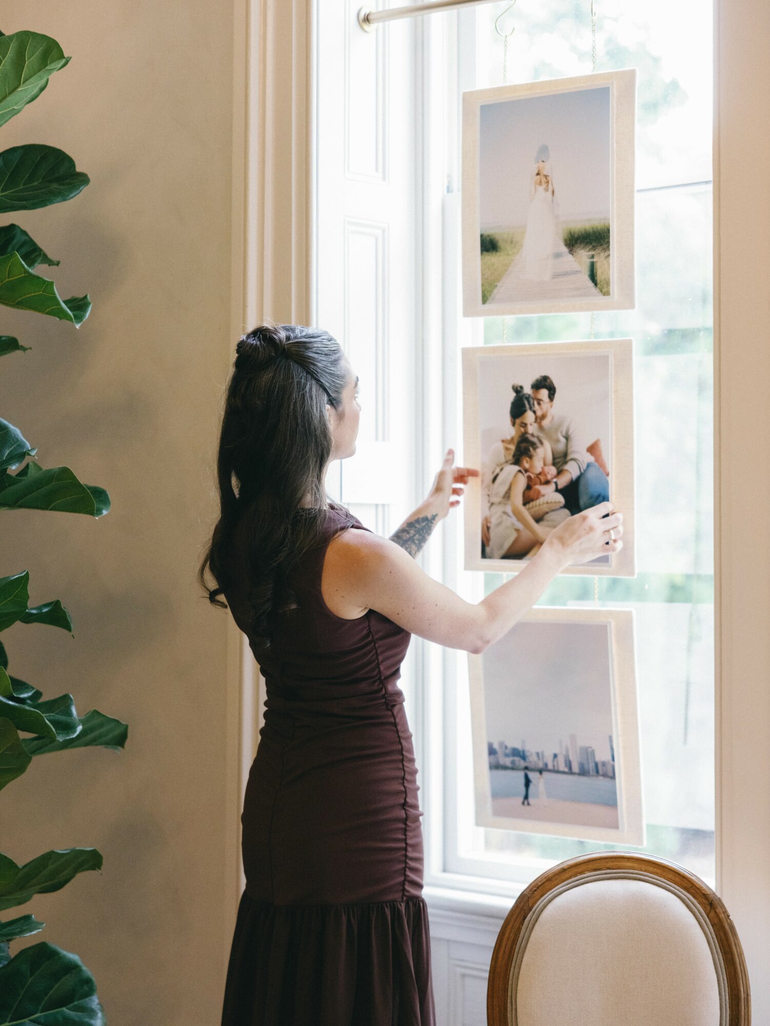 Woman arranging framed photographs on a window ledge, with a large window and a plant nearby.