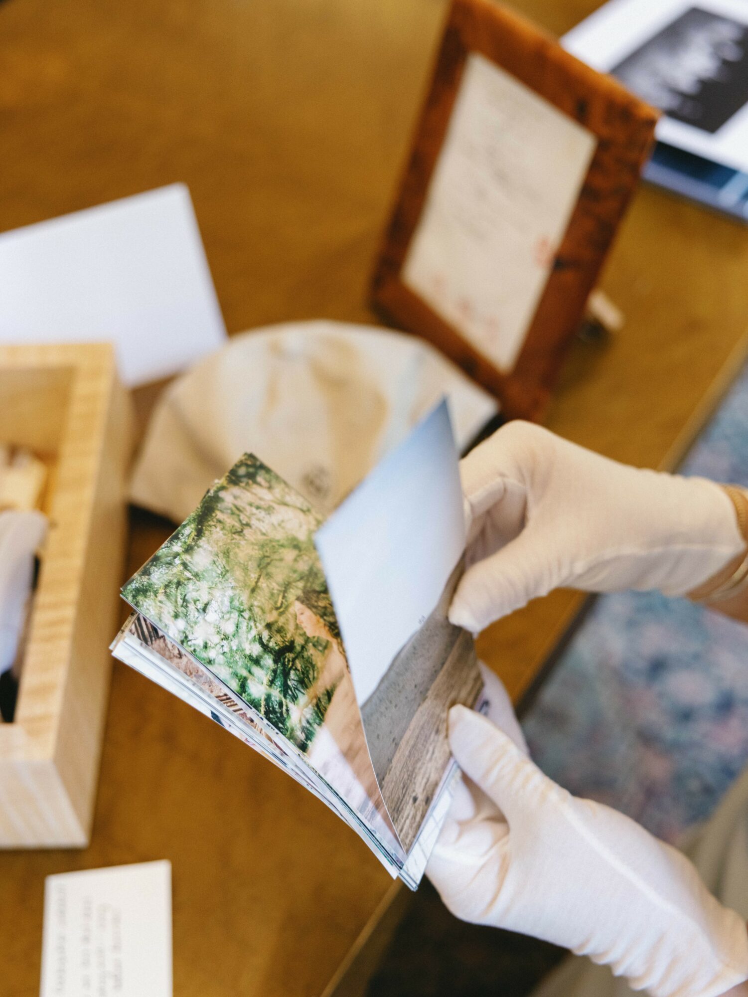 Person wearing white gloves flipping through a small photo album on a wooden table, with other items nearby.