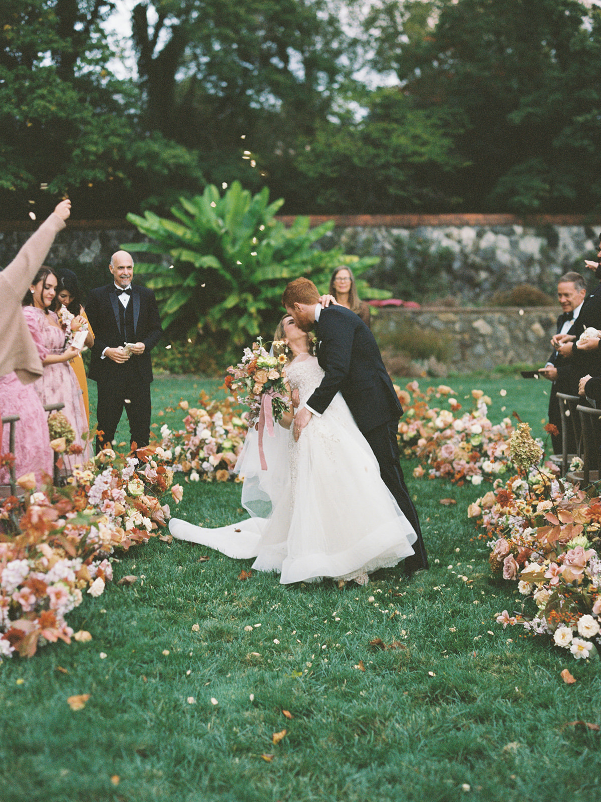 A bride and groom kiss in a garden with floral arrangements, surrounded by guests, trees, and a stone wall.