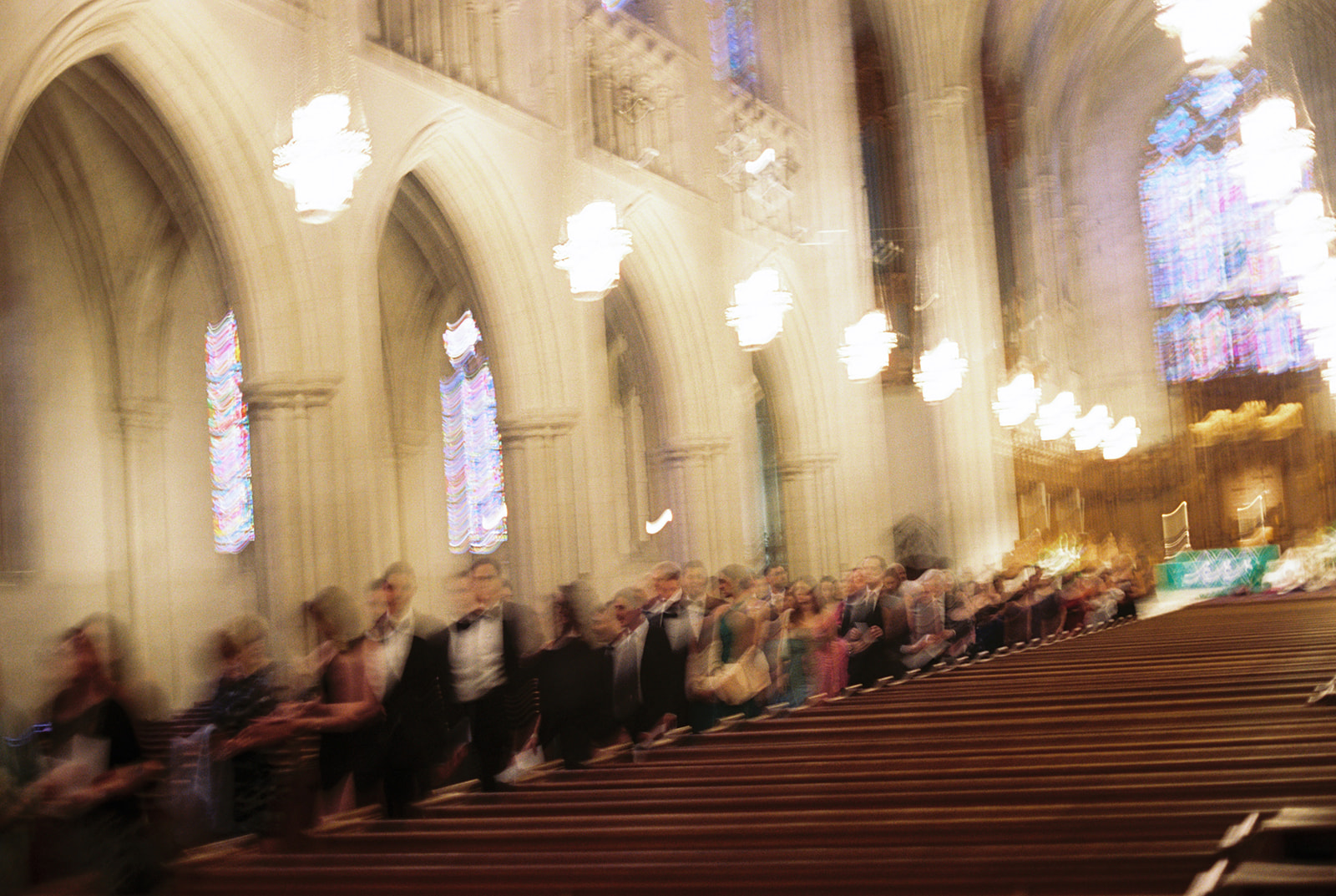Inside a church with high arches, stained glass windows, and rows of pews, with people standing in line.