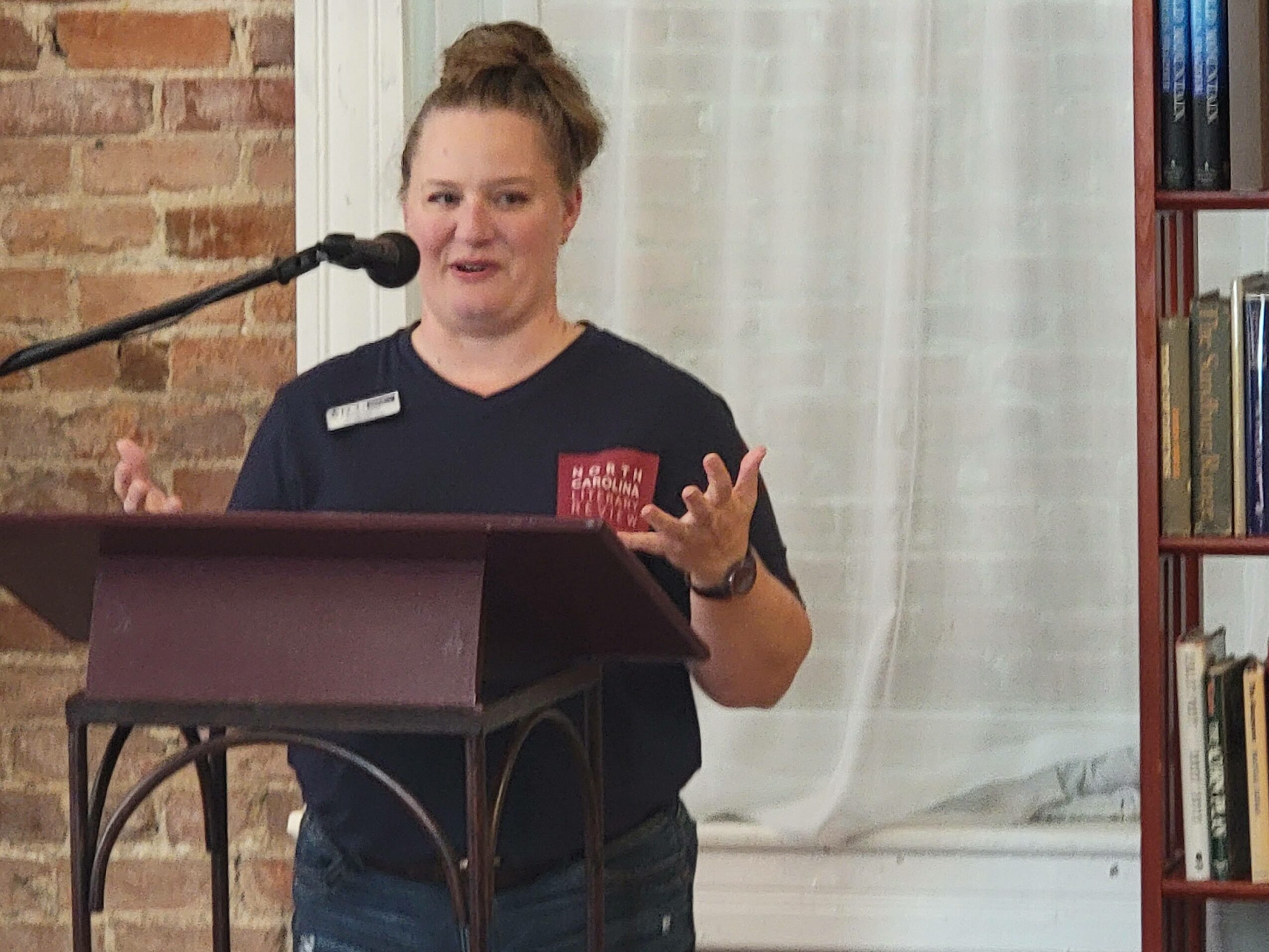 Woman speaking at a podium with a microphone, wearing a dark shirt with a name tag, in a room with a brick wall and bookshelf.