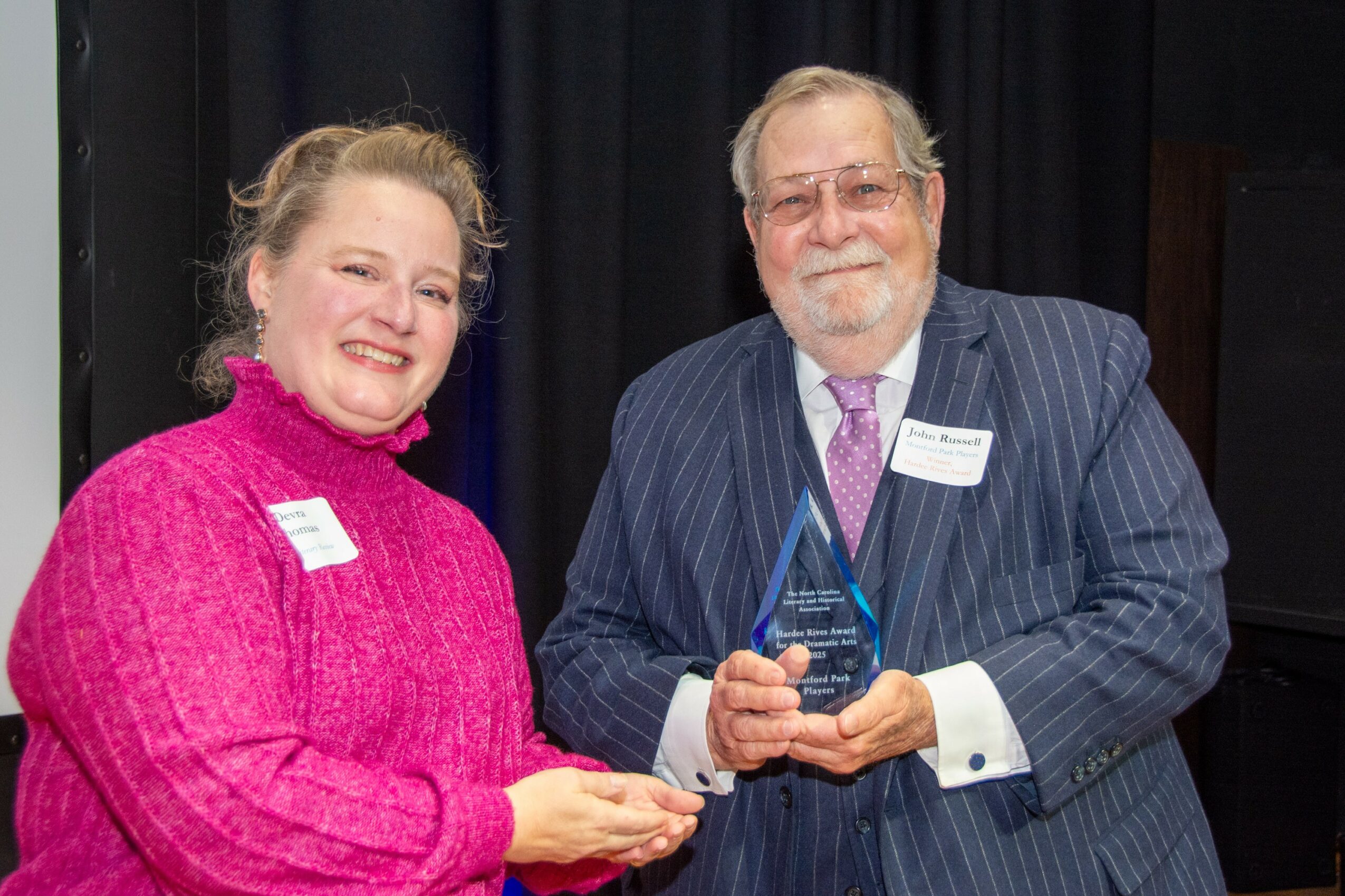 Two people smiling, one holding a glass award, standing in front of a dark curtain, dressed in formal attire.